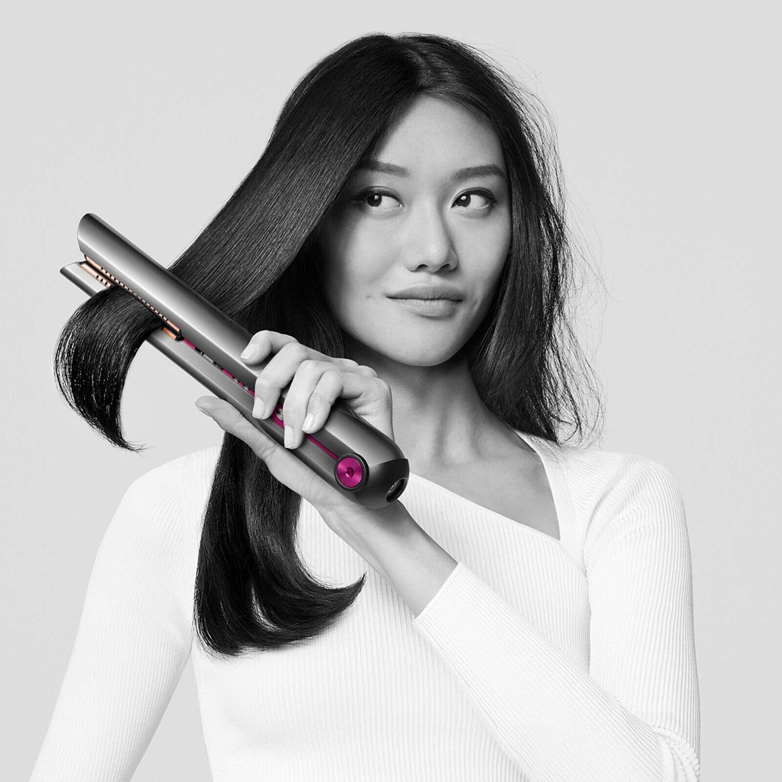 Frau benutzt graues Dyson-Haarglätteisen auf ihrem dunklen Haar, schaut in die Kamera.