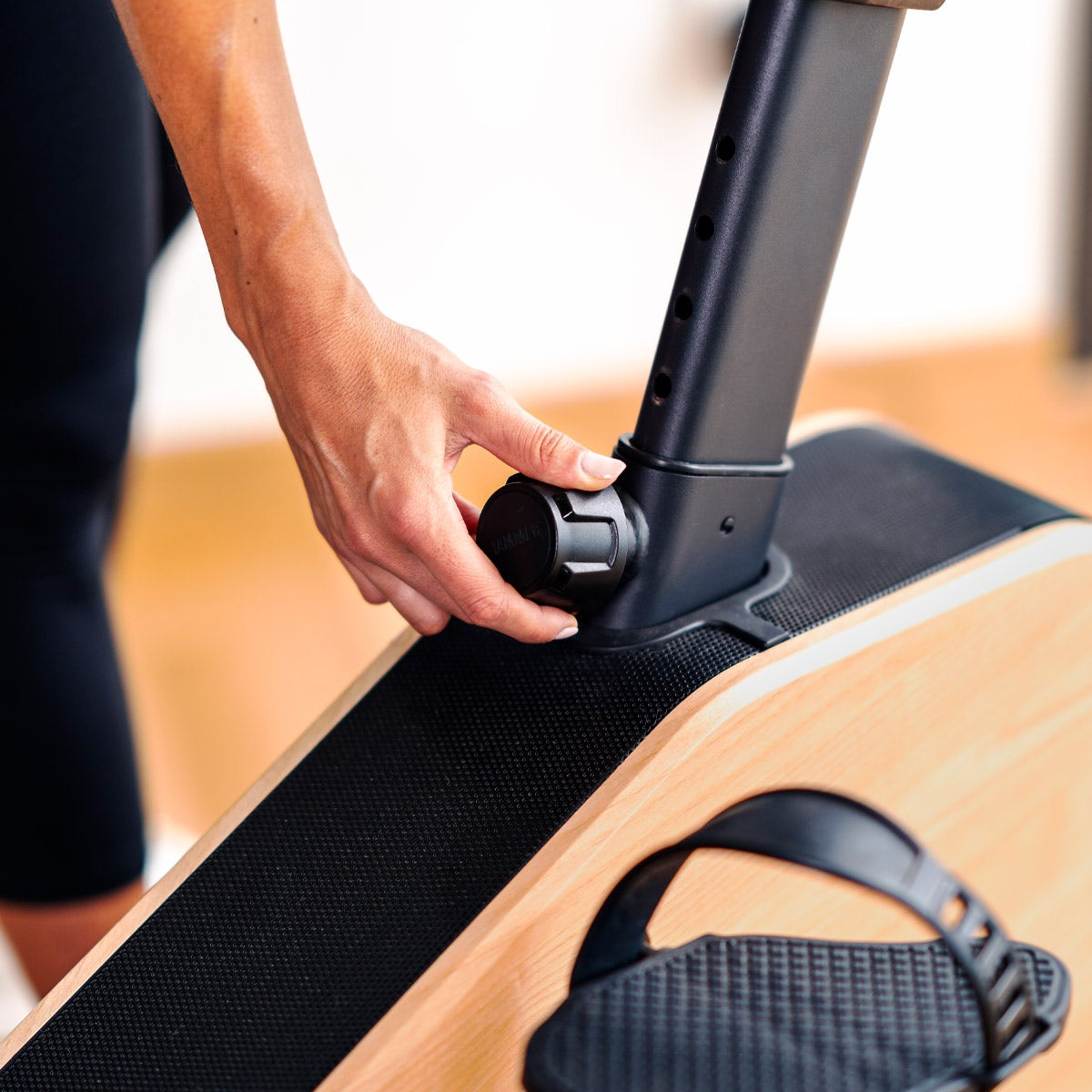 Person stellt schwarzen Knopf an einem hölzernen Trainingsgerät ein. Schwarze Beinbekleidung sichtbar.