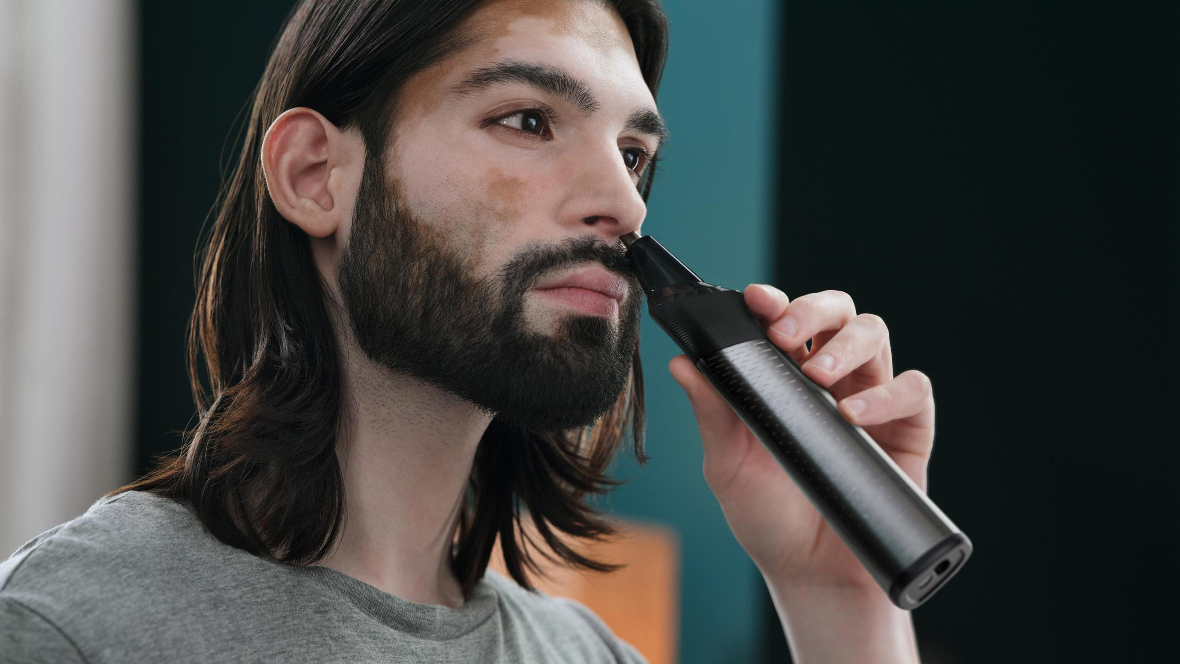 Mann, der seine Nasenhaare mit einem schwarzen Trimmer trimmt. Er hat lange dunkle Haare und einen Bart.