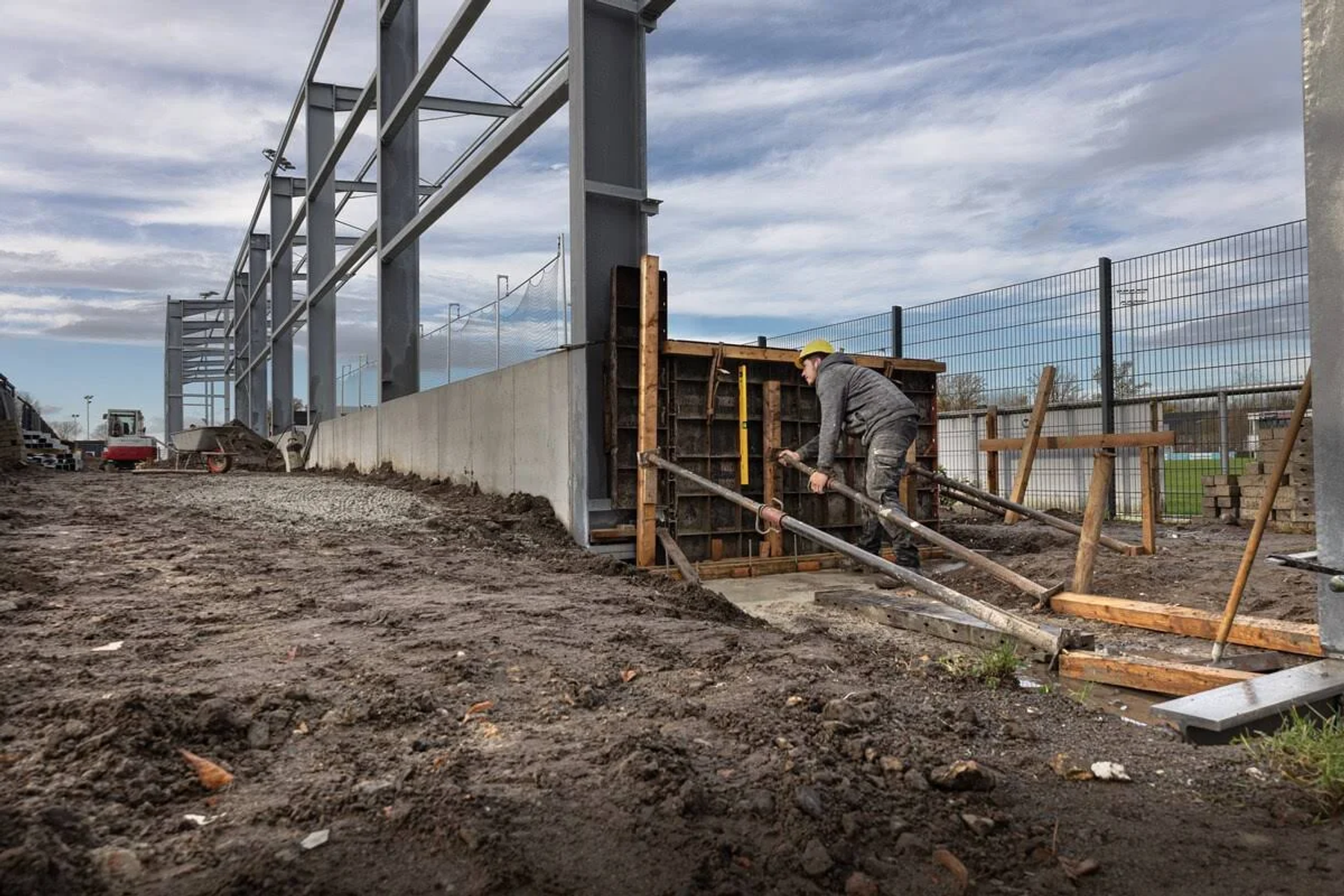 Baustelle mit Arbeiter, Holzform, Metallträgern und bewölktem Himmel im Hintergrund.