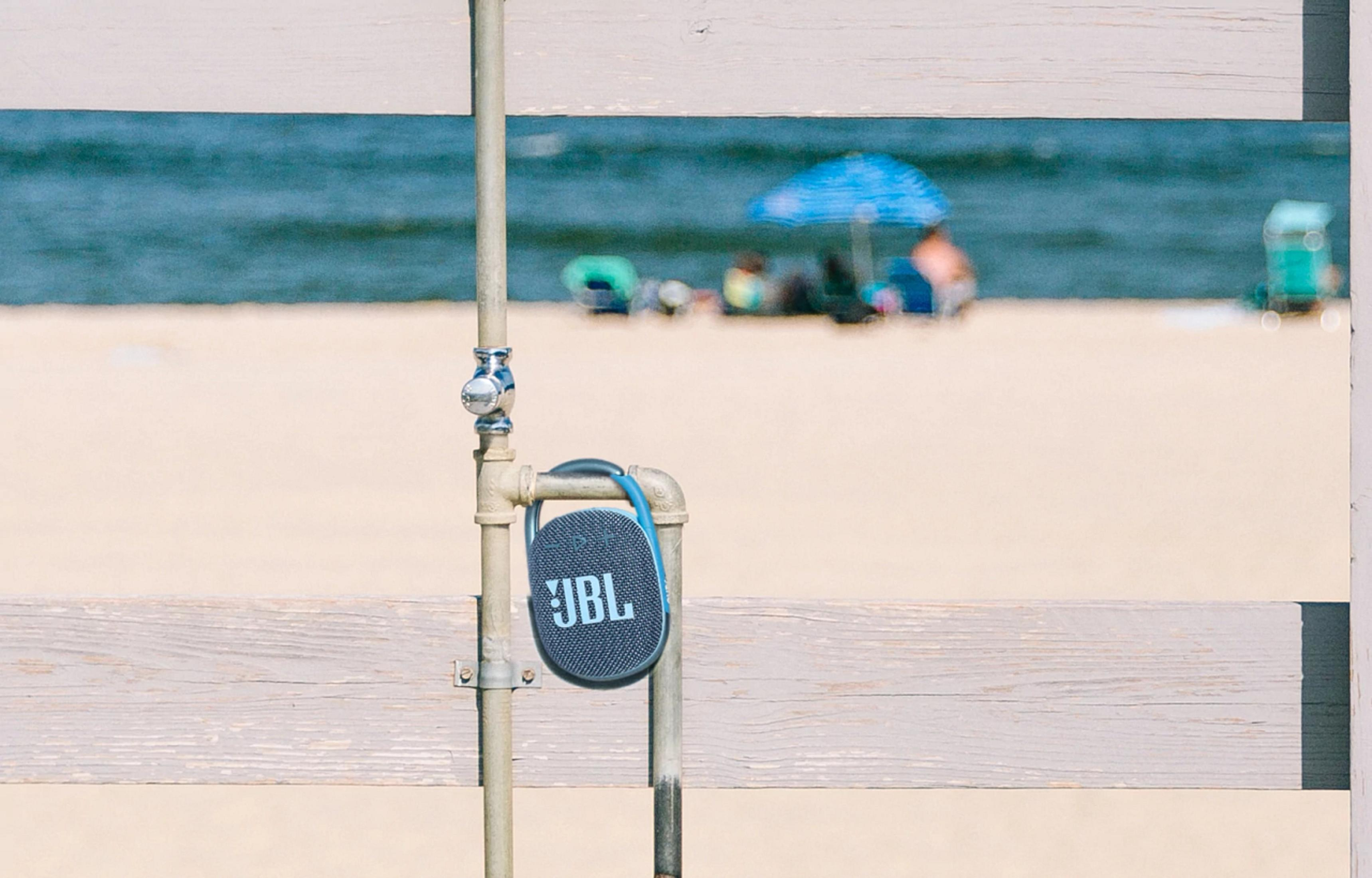 Głośnik JBL zawieszony na rurze na plaży, z ludźmi i morzem w tle.