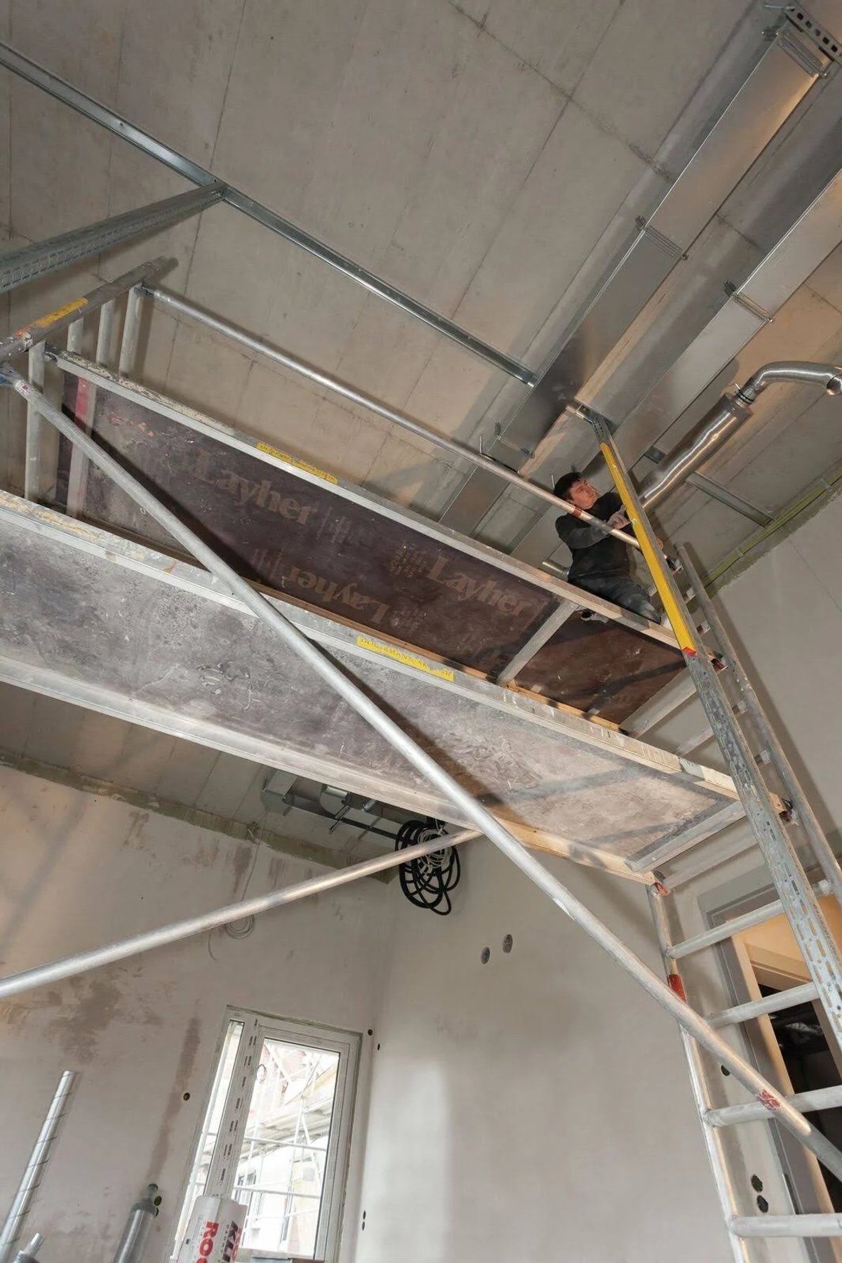 Arbeiter auf einem Gerüst in einem Raum, arbeitet an einer Decke mit Rohren.