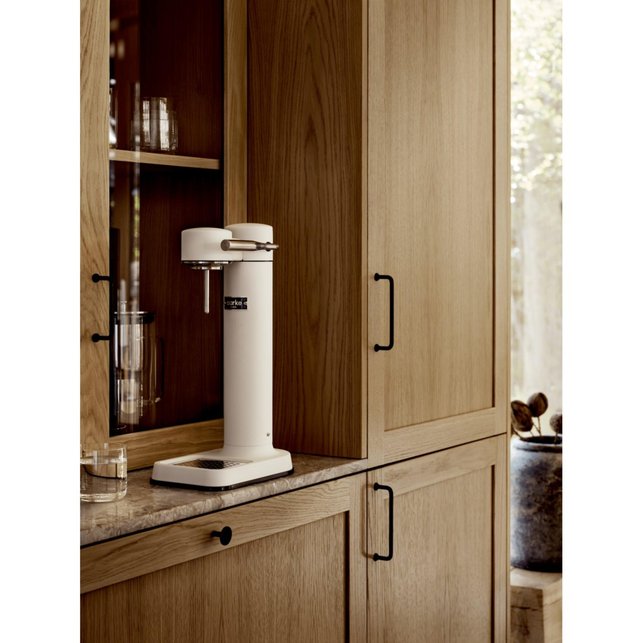 Un produttore di soda bianco su un piano di lavoro in legno, con armadi in legno sullo sfondo. C'è un bicchiere accanto.
