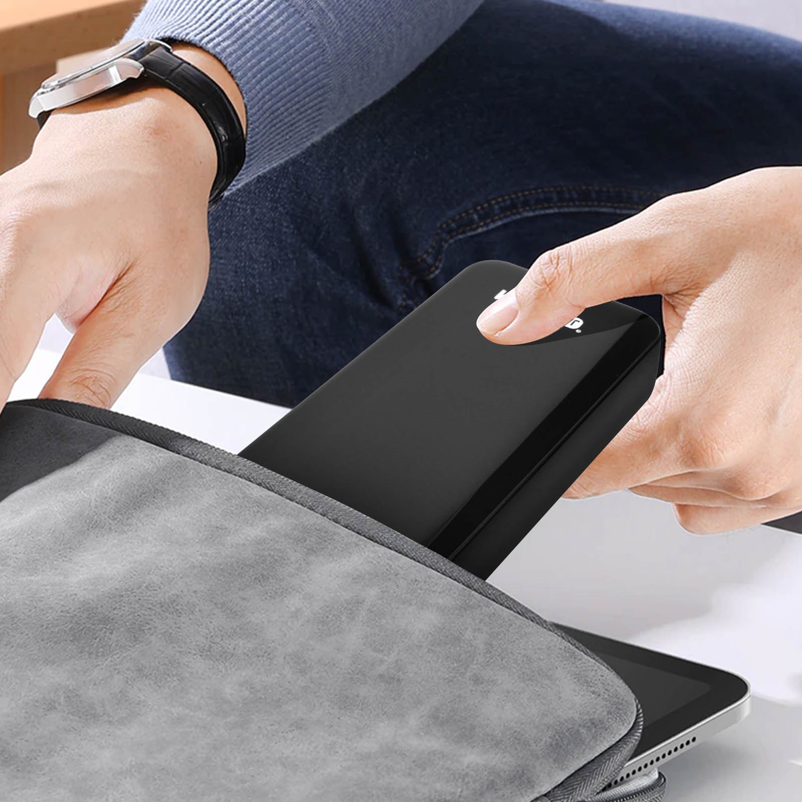 Eine Person legt ein schwarzes Gerät in eine graue Tasche. Eine Uhr am Handgelenk.
