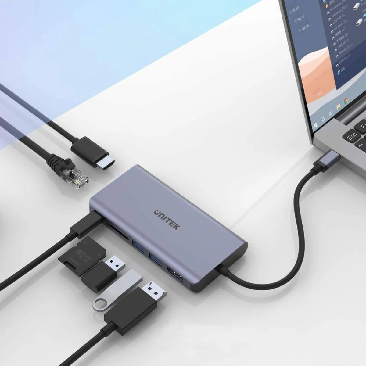 Ein grauer Hub mit USB-, Ethernet- und anderen Anschlüssen, der über Kabel an einen Laptop angeschlossen ist. Im Hintergrund ist ein Schreibtisch.