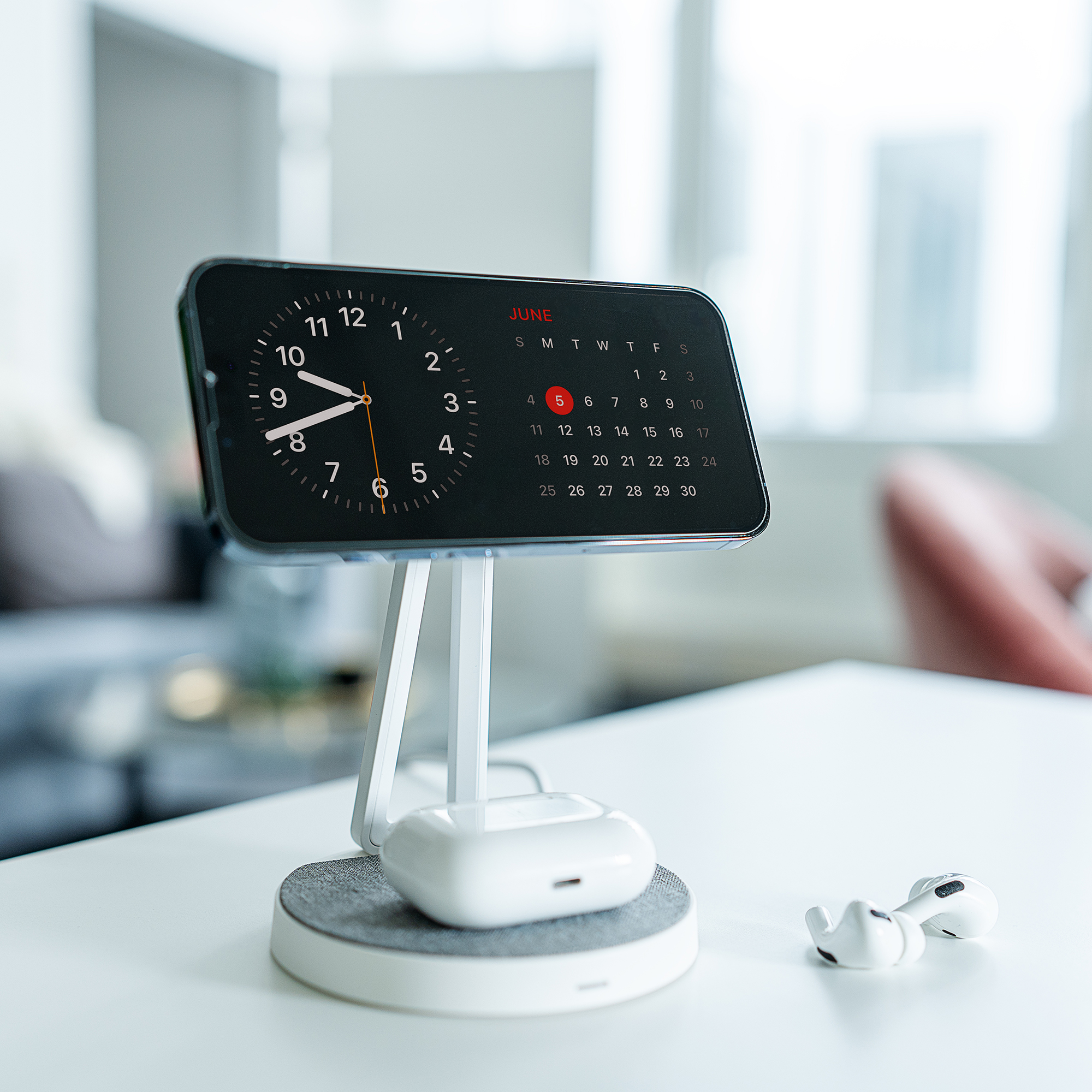 Telefon auf Ständer zeigt Zeit und Kalender. AirPods auf Ladegerät und lose. Weißer Tisch, Hintergrund verschwommen.