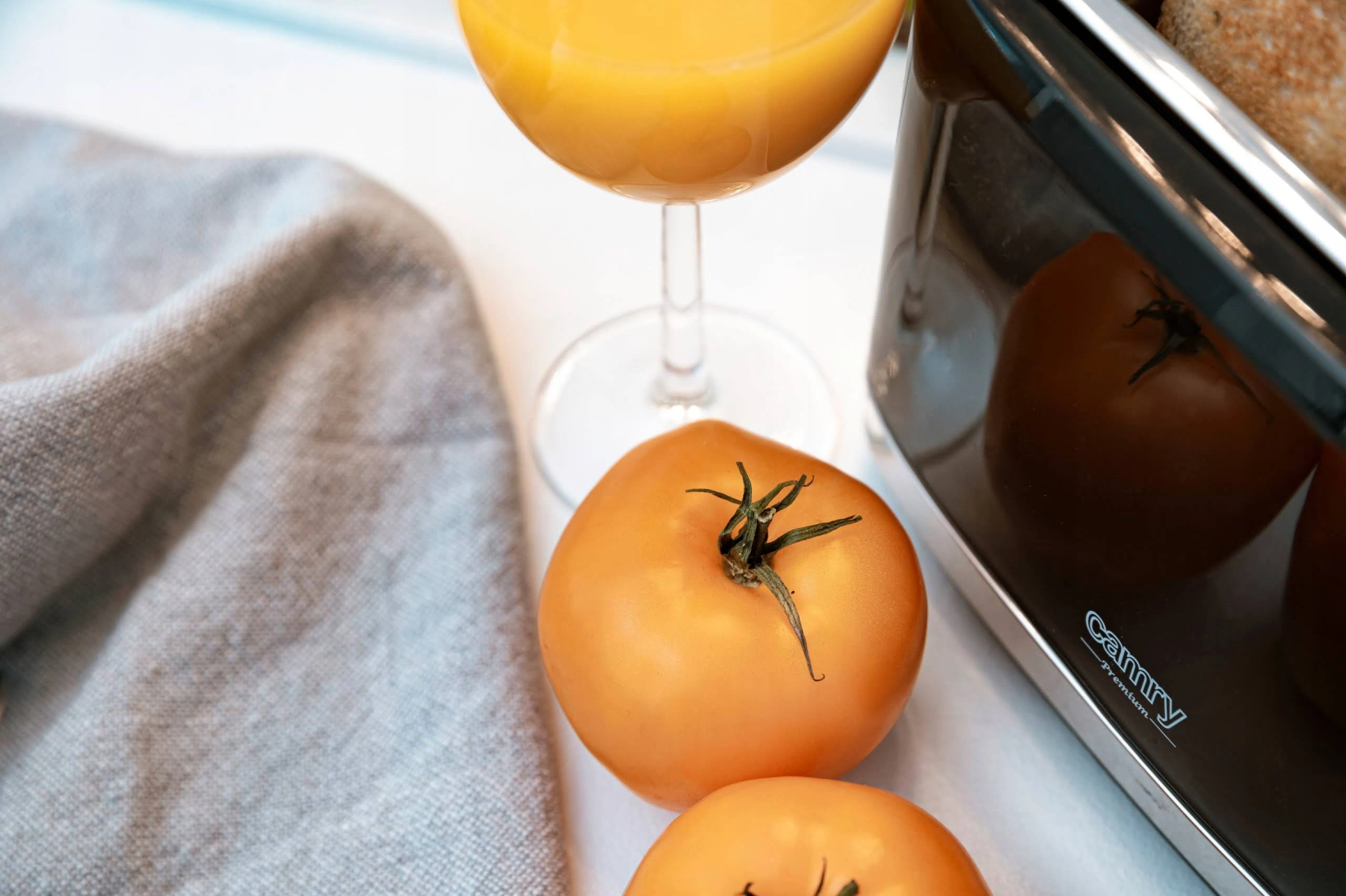 Sok pomarańczowy w szklance, pomidory i czarny toster na białej powierzchni.