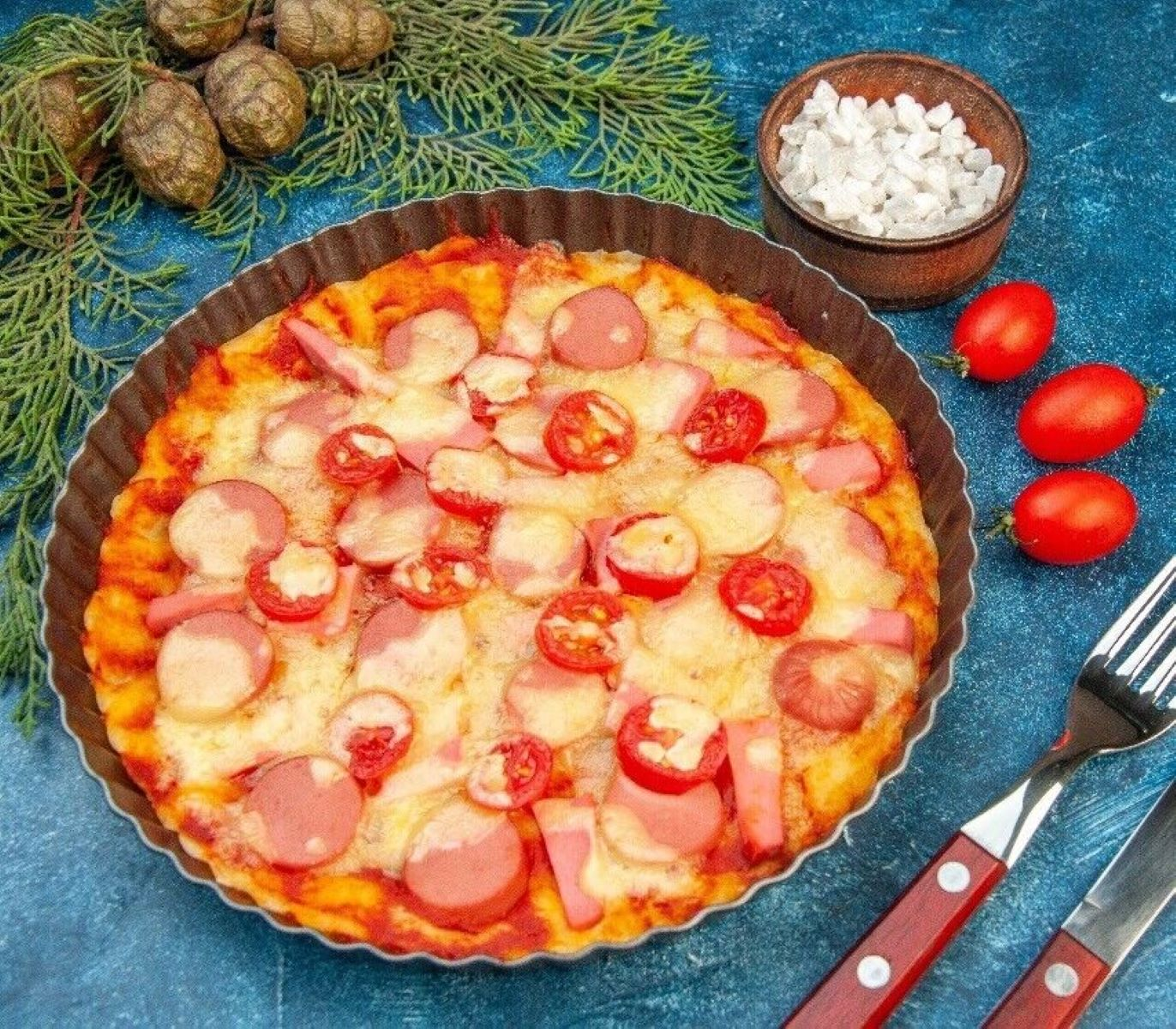 Pizza in einer Metallpfanne. Beläge sind Wurst, Tomaten und Käse auf einem blauen Tisch.