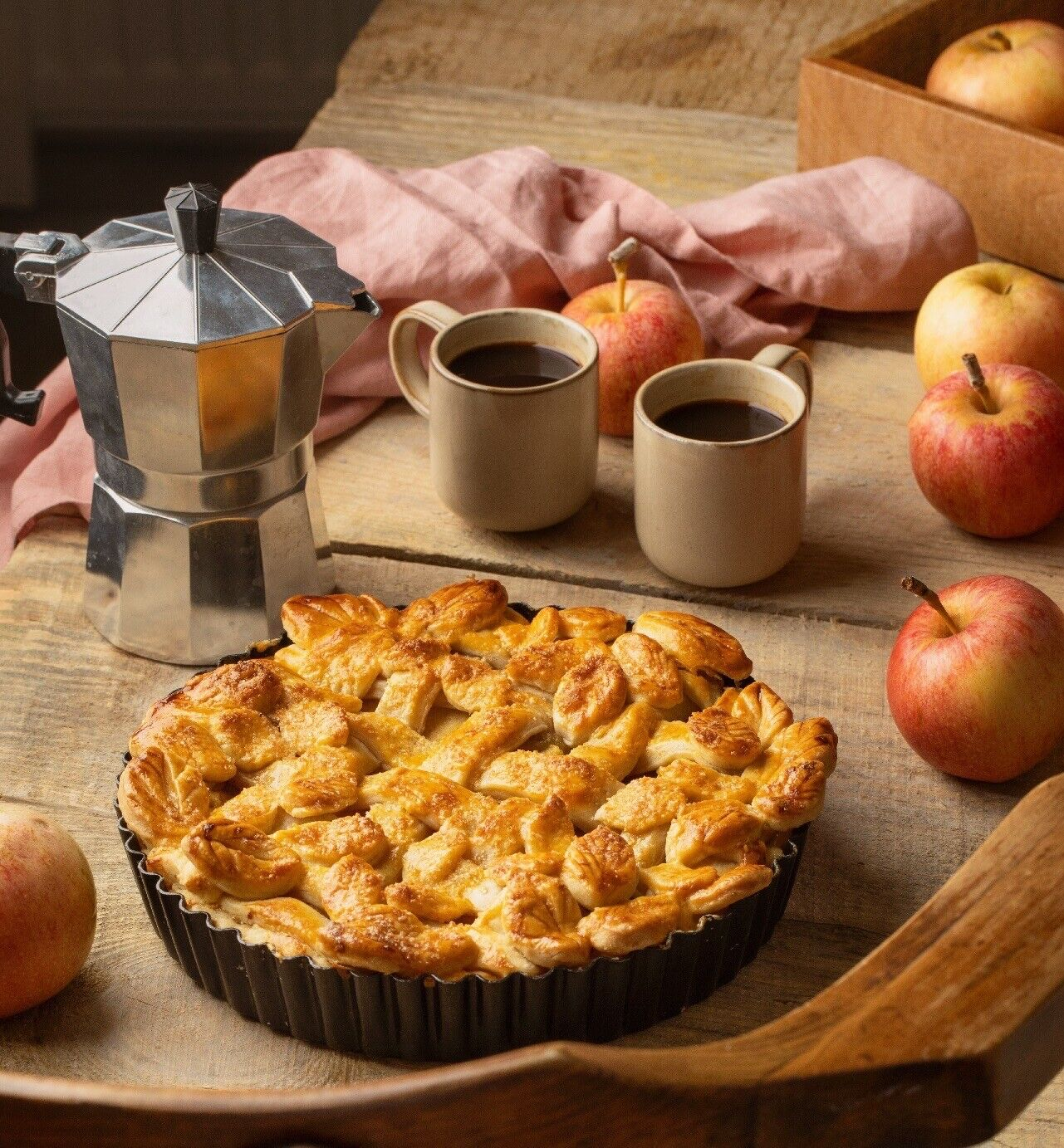 Apfelkuchen in einer Pfanne, Kaffeekanne, zwei Tassen Kaffee und Äpfel auf einem Holztisch.