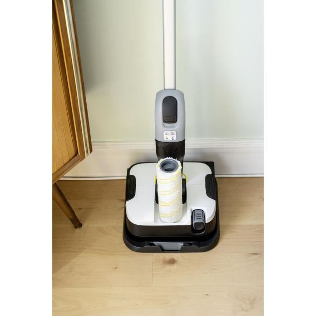 Un pulitore per pavimenti bianco e nero con un lungo manico. È su un pavimento in legno. Un piccolo mobile a sinistra.