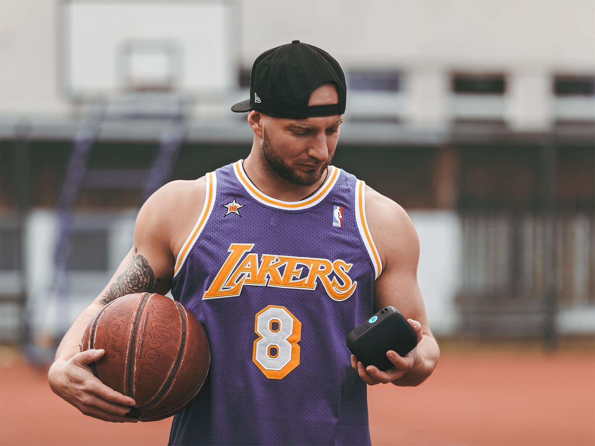 Mężczyzna w koszulce Lakers trzymający piłkę do koszykówki i małe czarne urządzenie. Tło boiska do koszykówki.