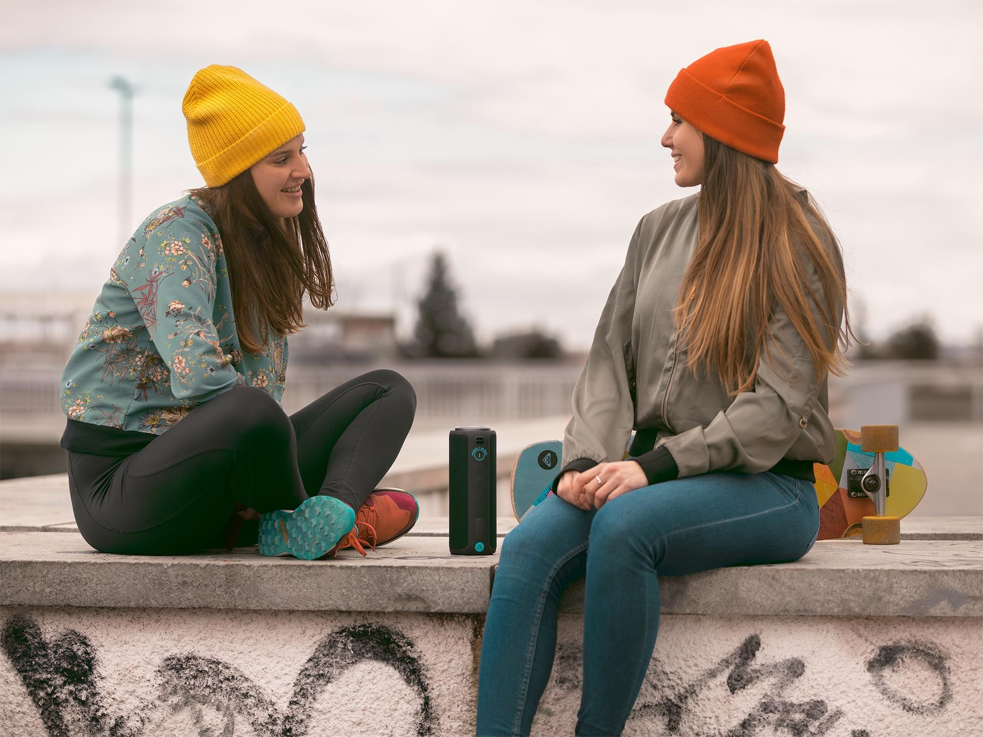 Dwie kobiety siedzące na betonowym murze, jedna w żółtej, a druga w pomarańczowej czapce, rozmawiają ze sobą.