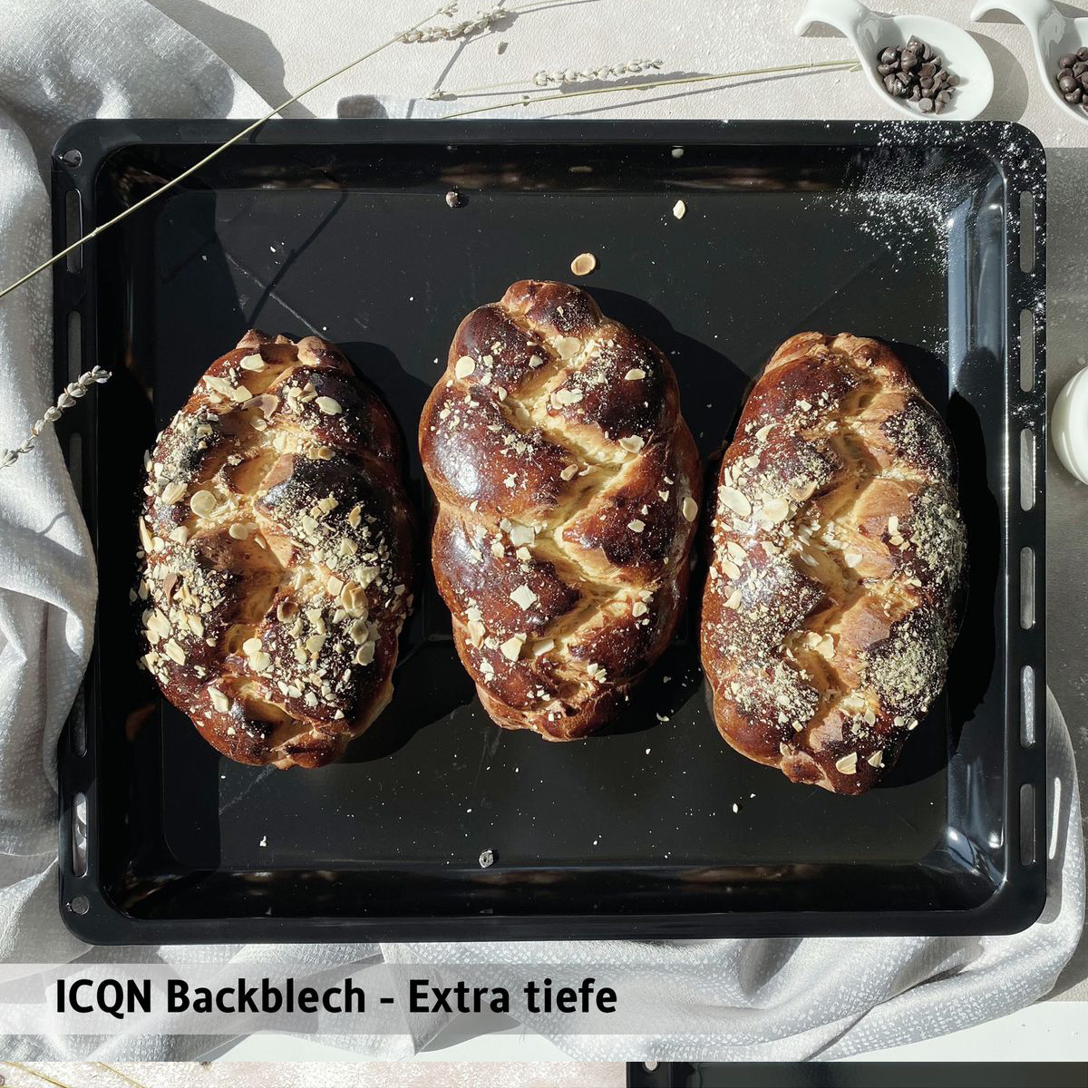 Drei geflochtene Brotlaibe mit Mandeln auf schwarzem Backblech, heller Hintergrund.