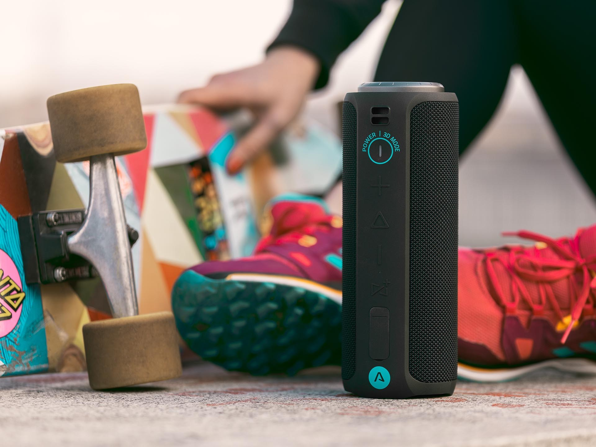 Czarny i turkusowy głośnik przenośny z deskorolką, butami i osobą w tle.