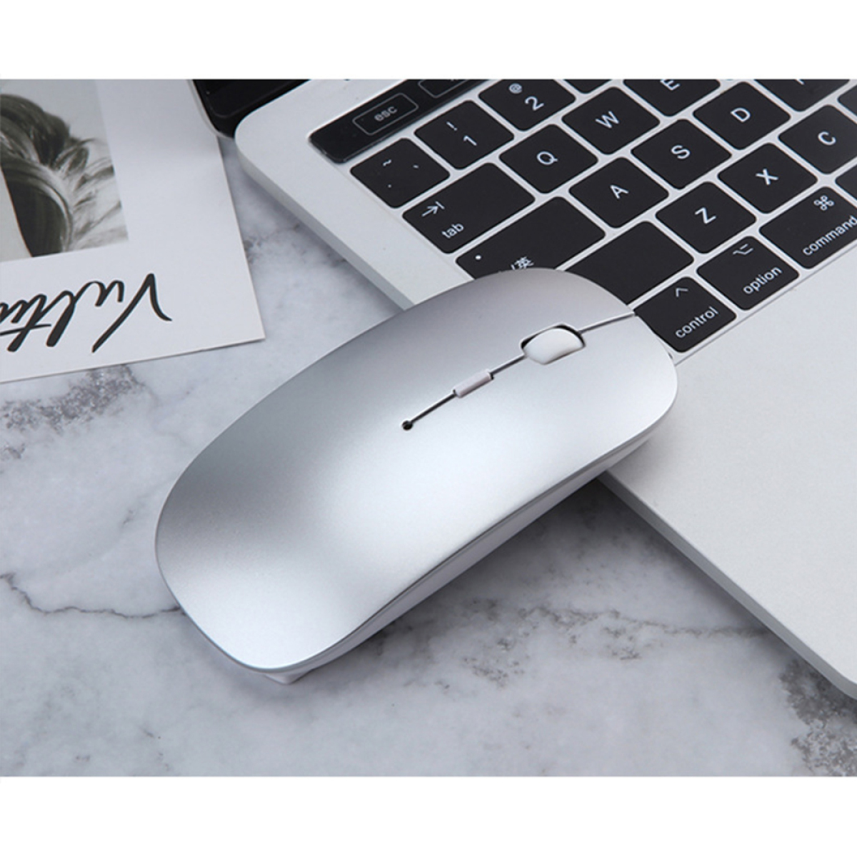 Eine silberne Maus liegt auf einer Laptop-Tastatur. Links liegt ein weißes Papier. Der Hintergrund ist Marmor.