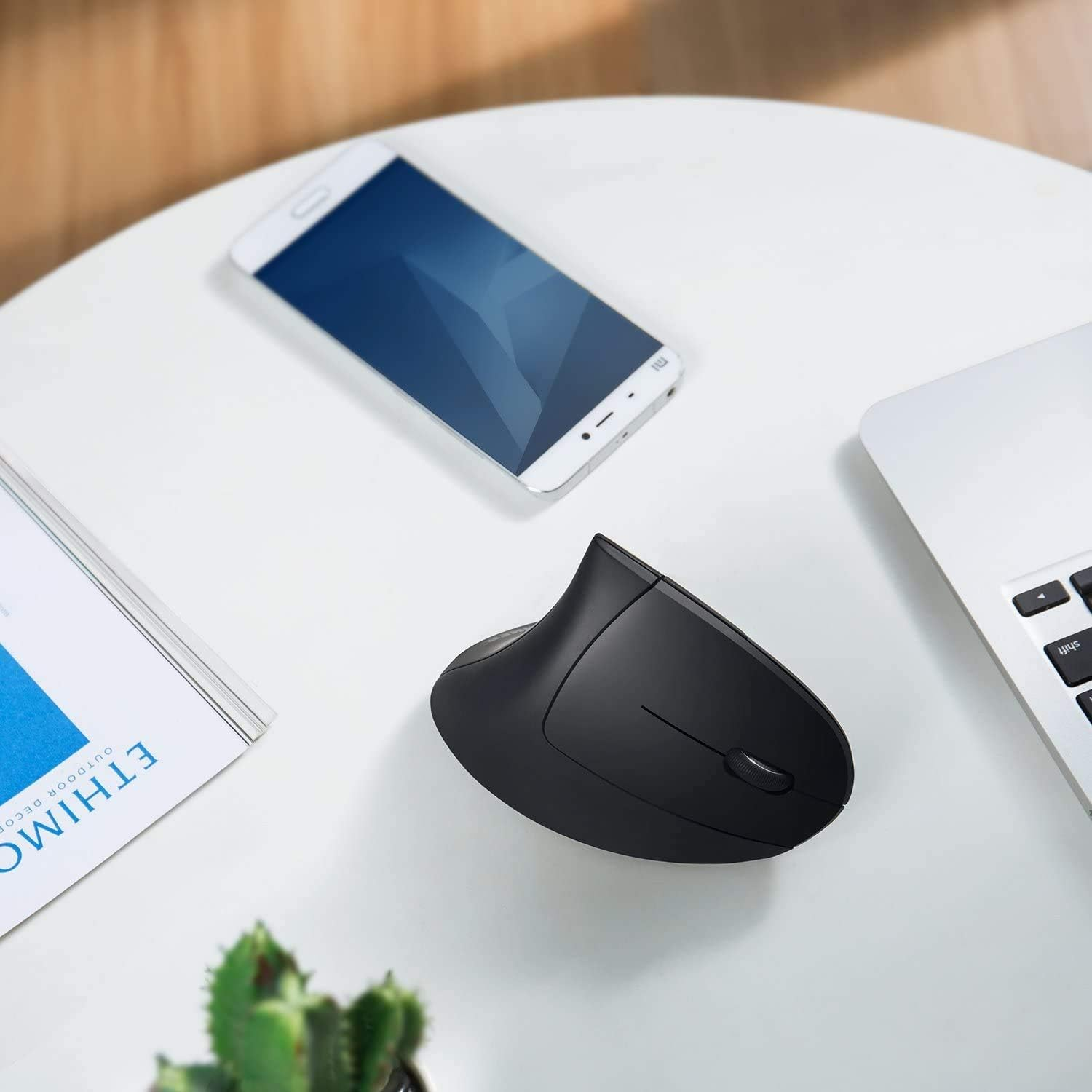 Schwarze ergonomische Maus, weißes Telefon und ein Laptop auf einem weißen Tisch. Links liegt ein Buch.