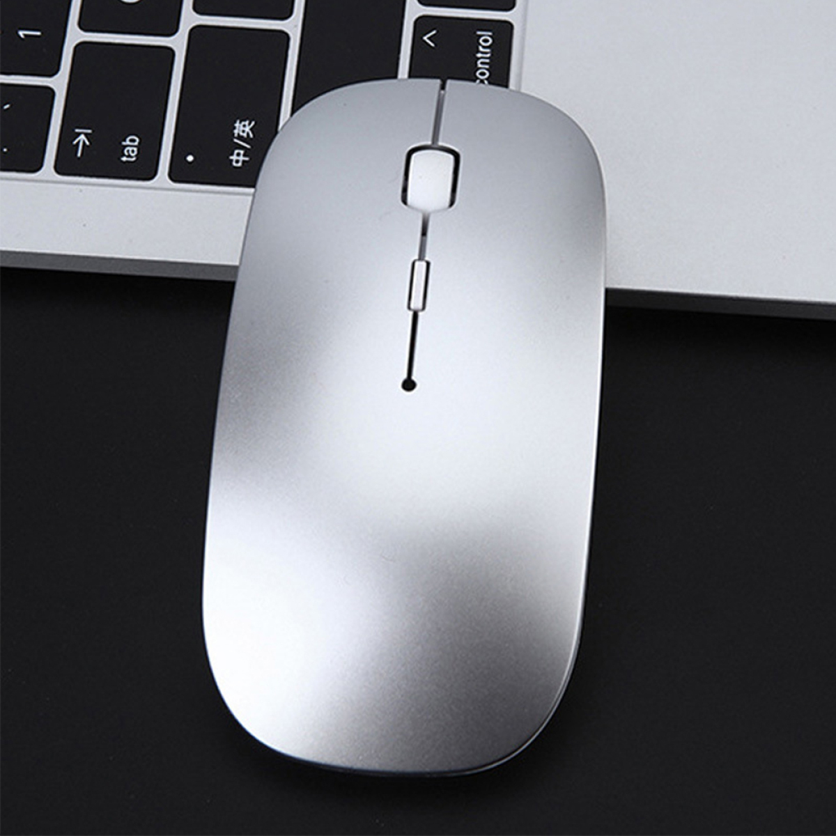 Eine silberne kabellose Maus auf einer schwarzen Oberfläche in der Nähe einer Tastatur. Die Maus ist schlank mit zwei Tasten und einem Scrollrad.