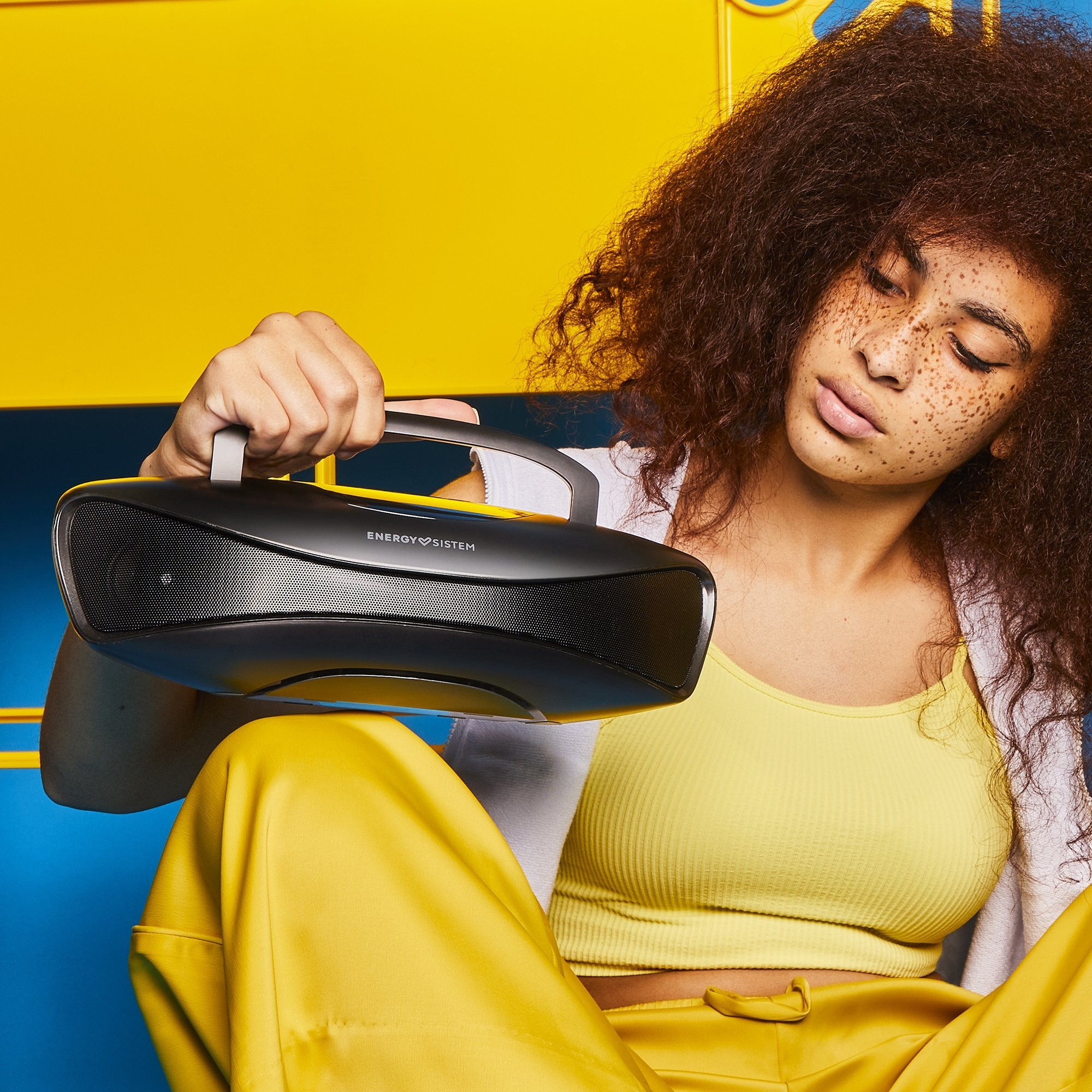 Una mujer sostiene un altavoz negro, se ve un fondo azul y amarillo.