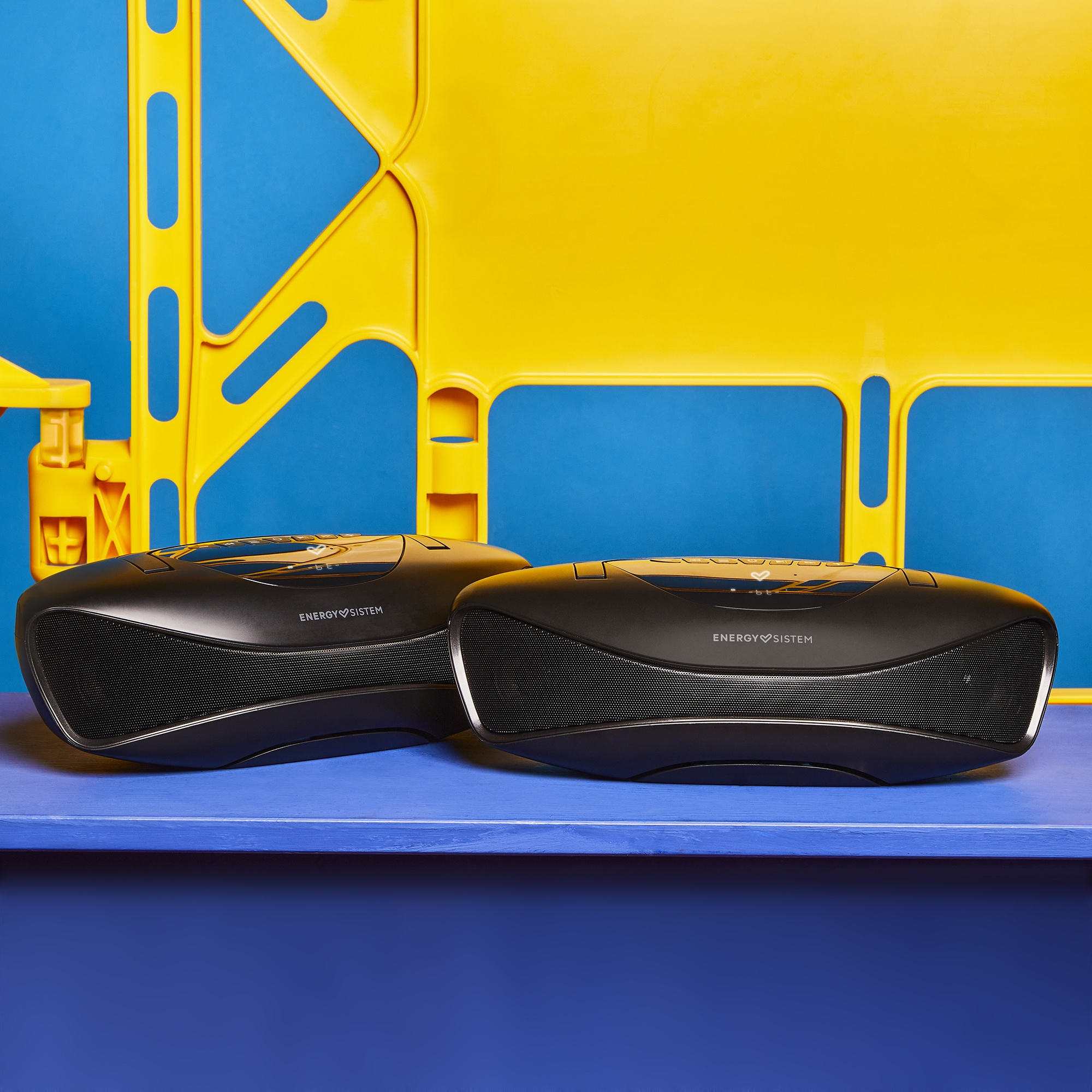 Dos altavoces boombox negros sobre una mesa azul con fondo amarillo. Los altavoces tienen un frente de malla.