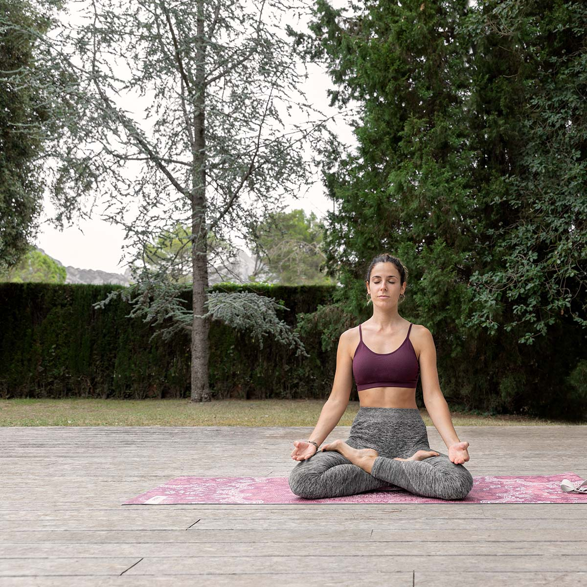 Mujer en pose de yoga en una esterilla en un parque. Árboles verdes, terraza de madera, cielo nublado.
