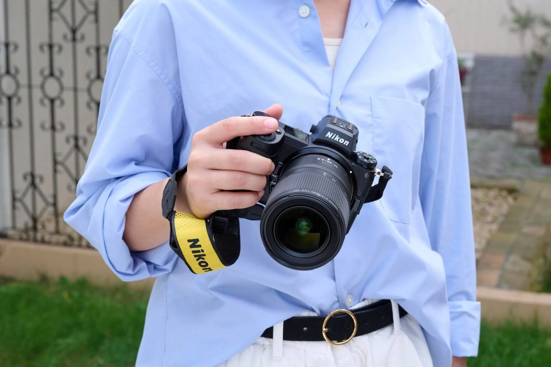 Persona sosteniendo una cámara Nikon con lente. Correa amarilla. Camisa azul. Cinturón negro.