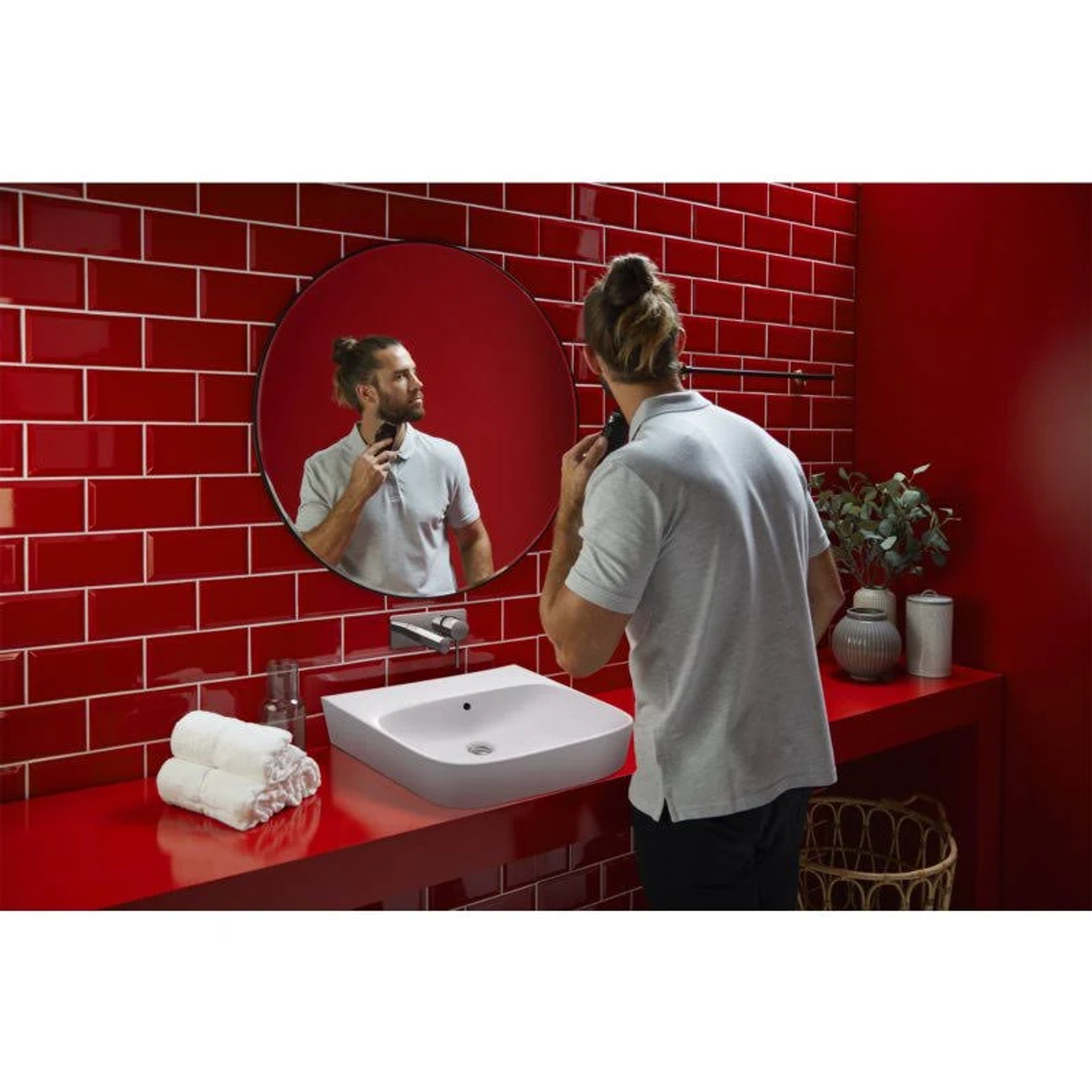 Ein Mann rasiert sich in einem rot gefliesten Badezimmer, blickt in einen runden Spiegel. Weißes Waschbecken, Handtücher und Pflanzen.