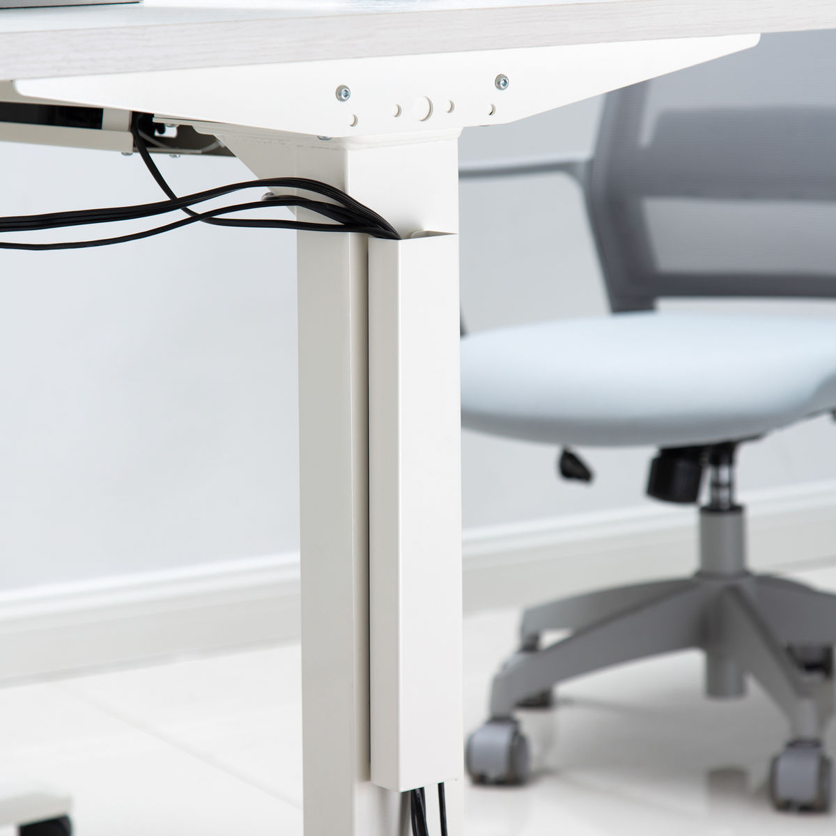 Escritorio y silla blancos. Cables negros cuelgan del escritorio. Silla gris claro con ruedas.