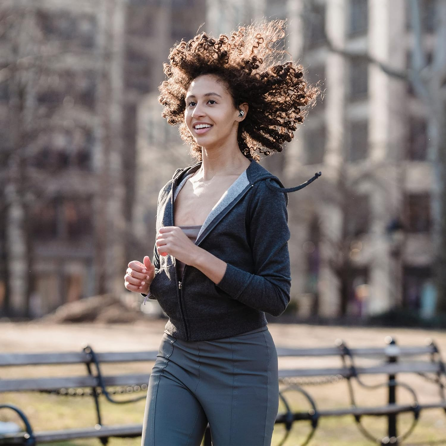 Una donna corre in un parco con una felpa grigia, pantaloni grigi e auricolari wireless.