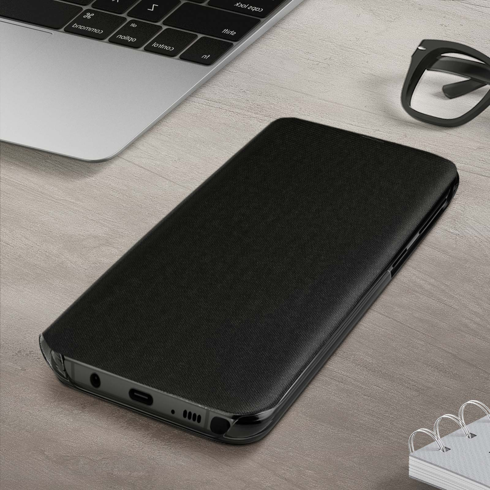 Un telefono nero in una custodia nera su un tavolo di legno. Laptop e occhiali sullo sfondo.