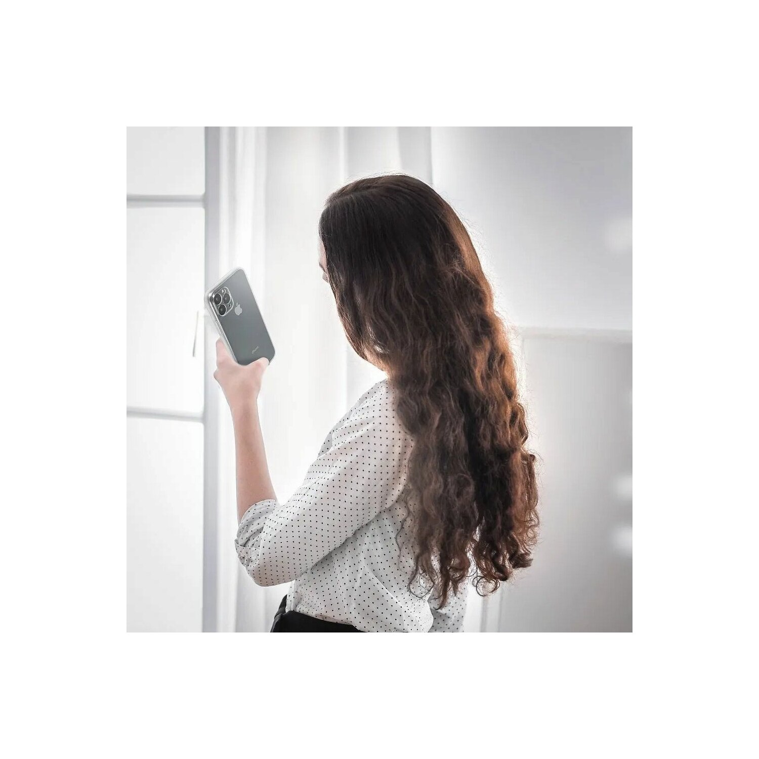 Frau hält Telefon, schaut auf den Bildschirm, langes welliges Haar, in der Nähe eines Fensters.