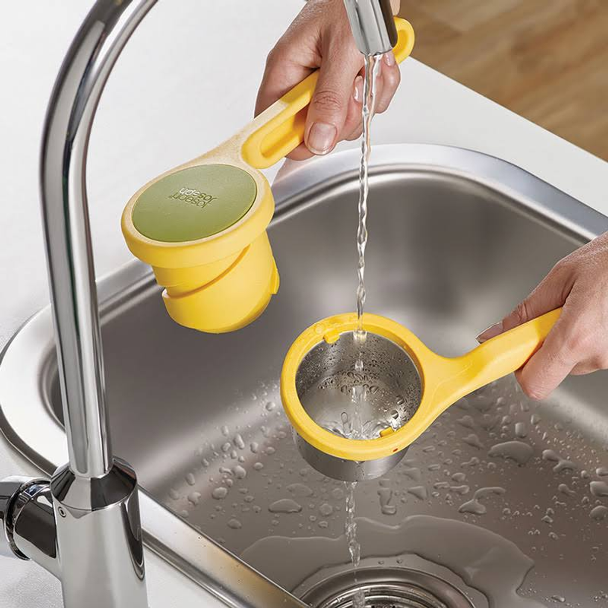 Mani che lavano utensili da cucina gialli e verdi. L'acqua scorre in un lavandino in acciaio inossidabile. Utensili in uso.