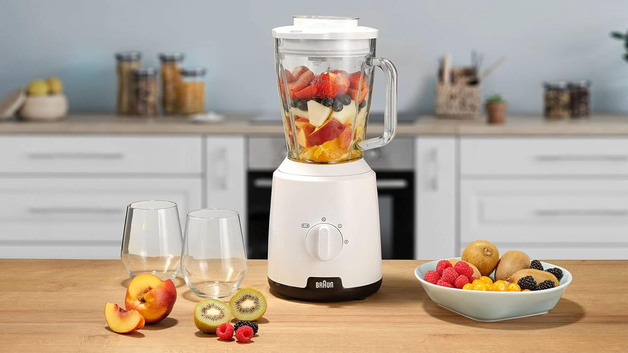 Batidora con fruta dentro, dos vasos y frutas en la mesa. Cocina al fondo.
