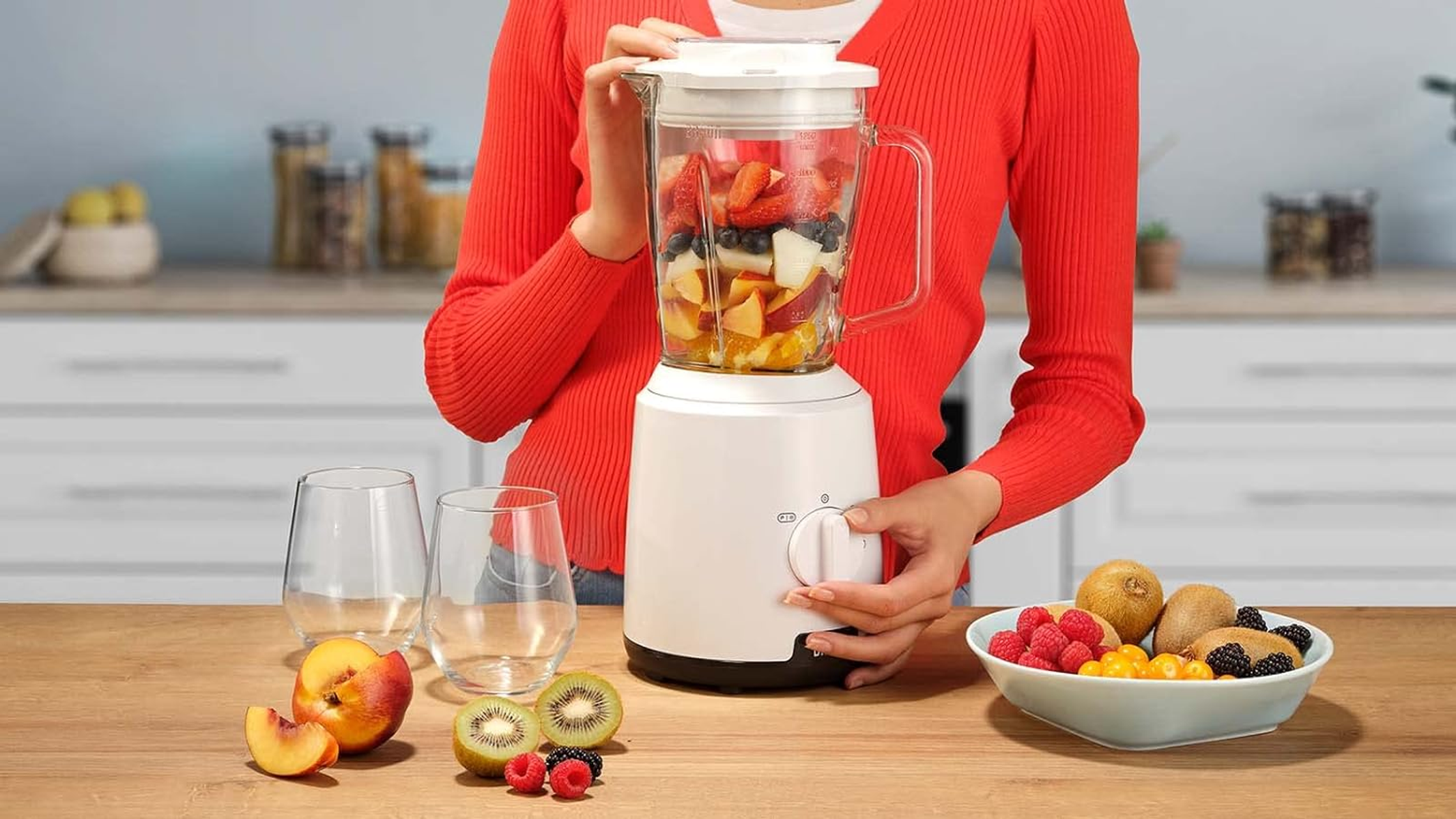 Mujer mezclando frutas en una licuadora. Hay frutas en la mesa y dos vasos vacíos.