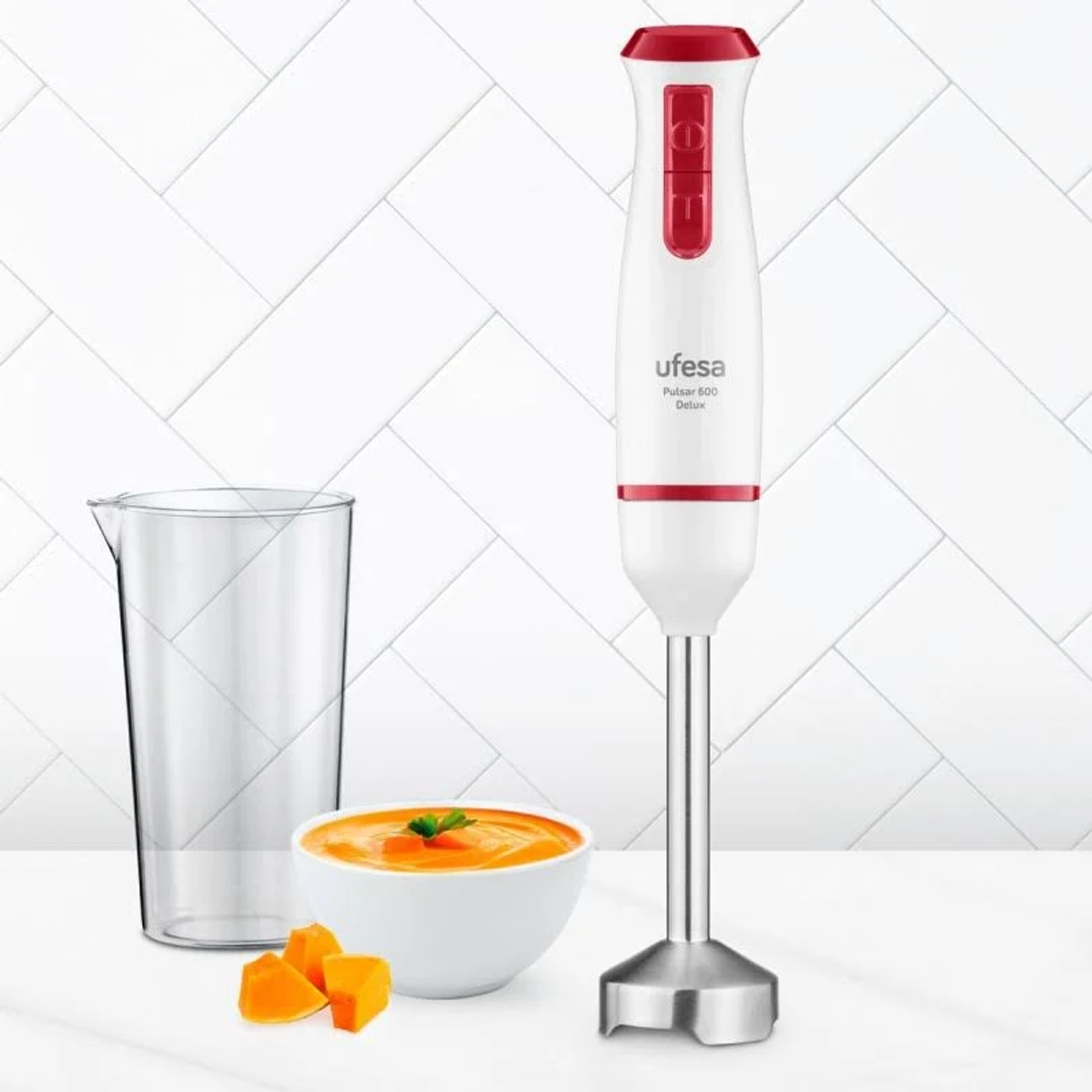Ein Ufesa-Stabmixer mit einer Schüssel Suppe, Kürbisstücken und einem Krug. Der Mixer ist weiß und rot.