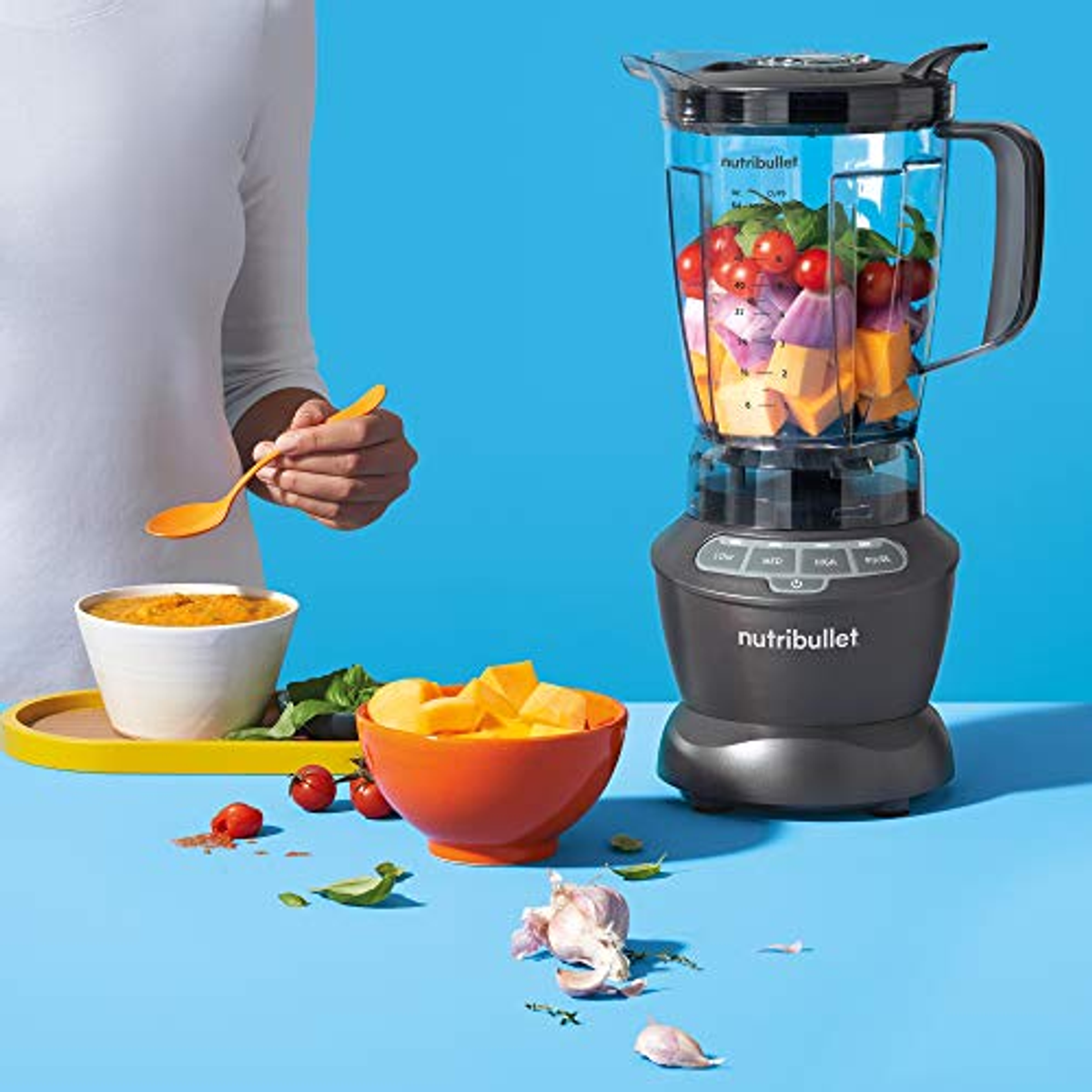 Licuadora con ingredientes: tomates, calabaza y ajo. Tazón de puré y cuchara. Fondo azul.