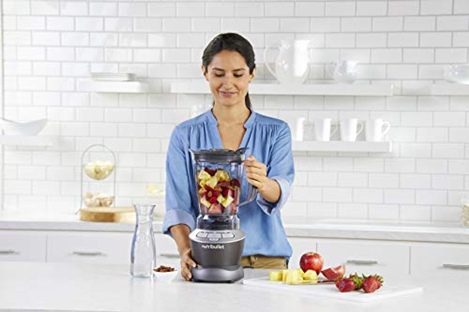 Mujer en la cocina usa una licuadora con manzanas y frutos rojos. Fondo de azulejos blancos.
