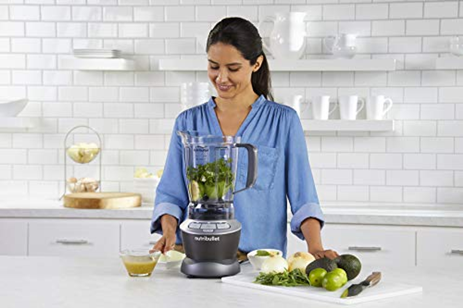 Una mujer usando una licuadora con ingredientes en una encimera de cocina. Hay frutas y verduras.