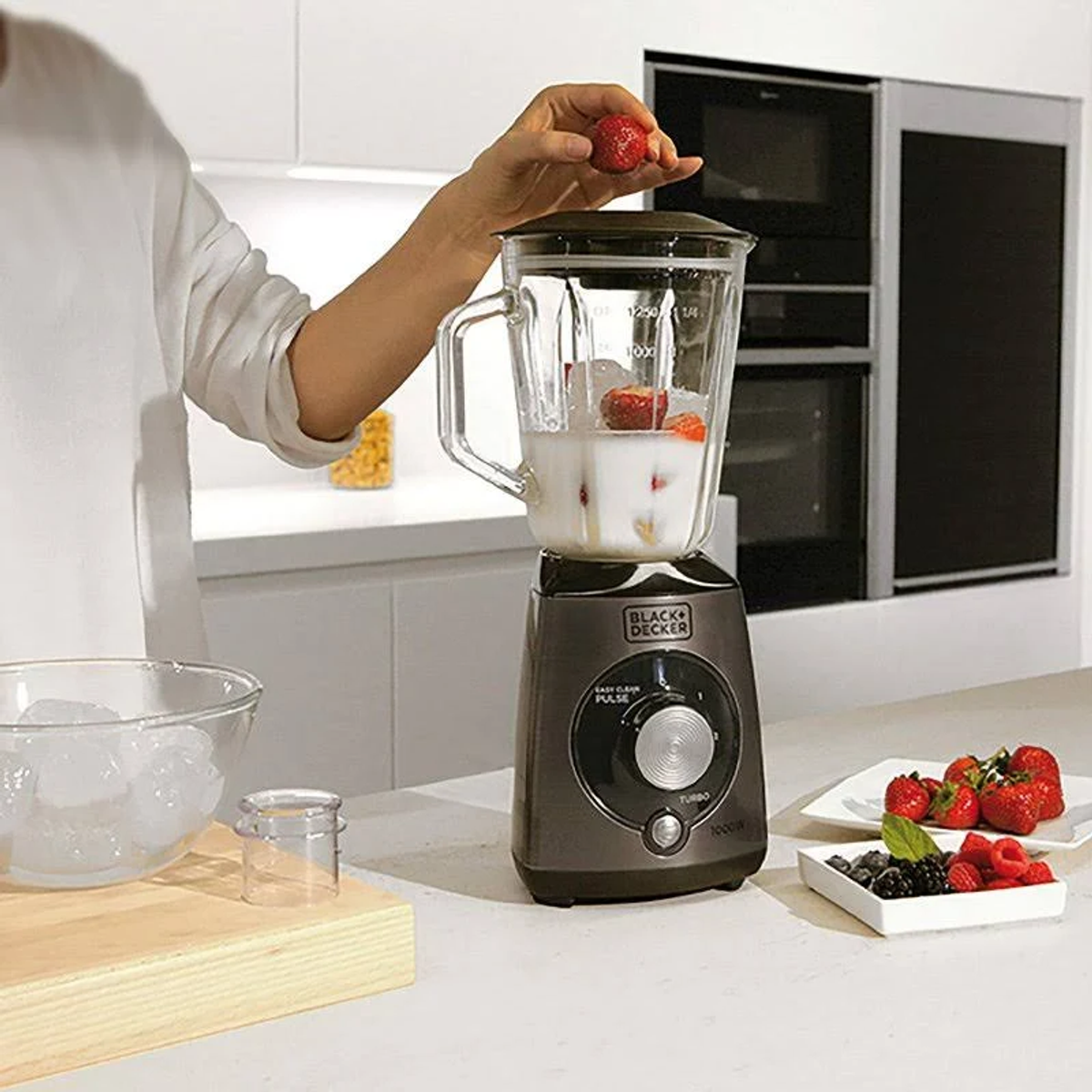 Osoba dodaje truskawkę do blendera z lodem i mlekiem. Inne owoce są na talerzu.