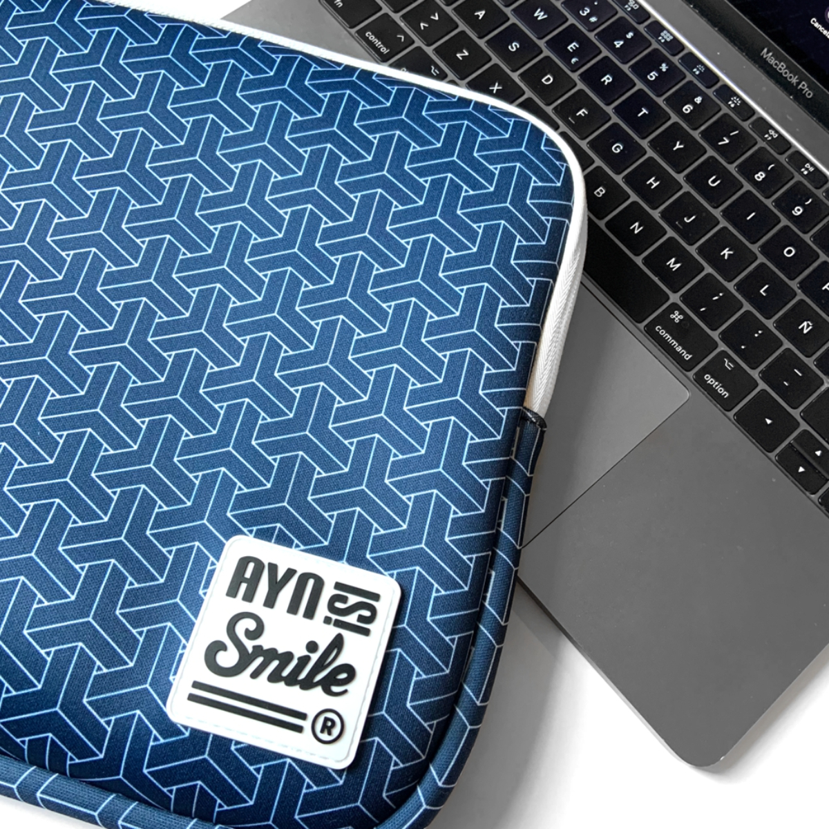 Funda para portátil con diseño geométrico azul y el logotipo de la marca, junto a un Macbook.
