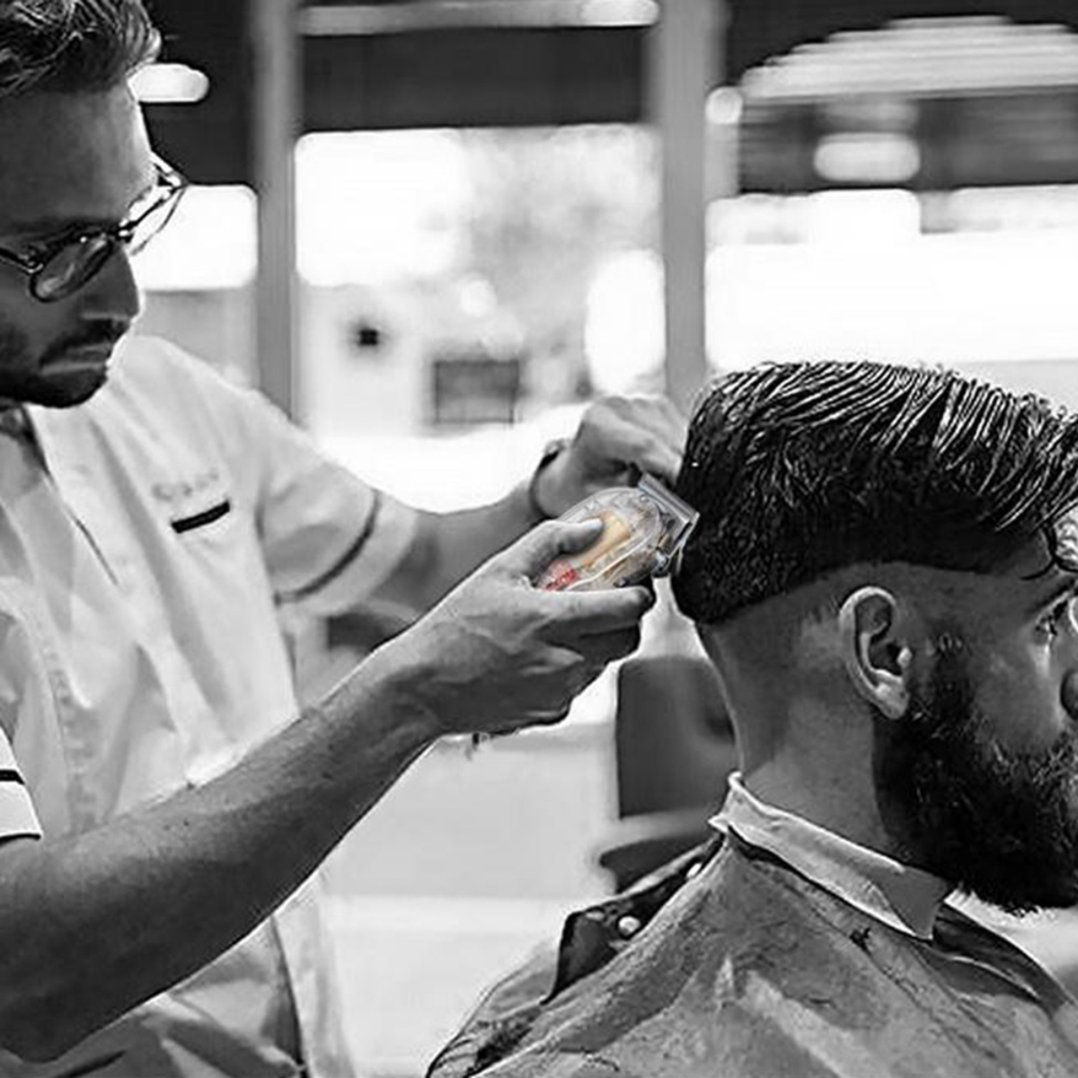 Barbero cortando el pelo. Hombre con camisa blanca, otro con capa de barbero. Foto en blanco y negro.