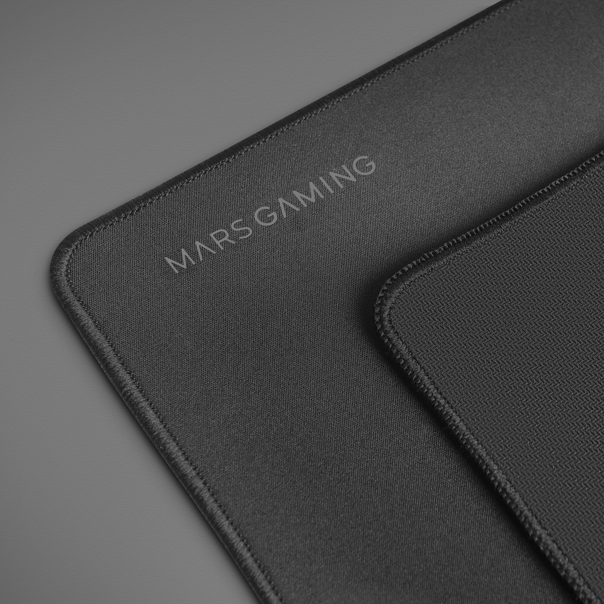 Zbliżenie dwóch czarnych podkładek pod mysz do gier na szarej powierzchni. Na jednej napis „MARS GAMING”.