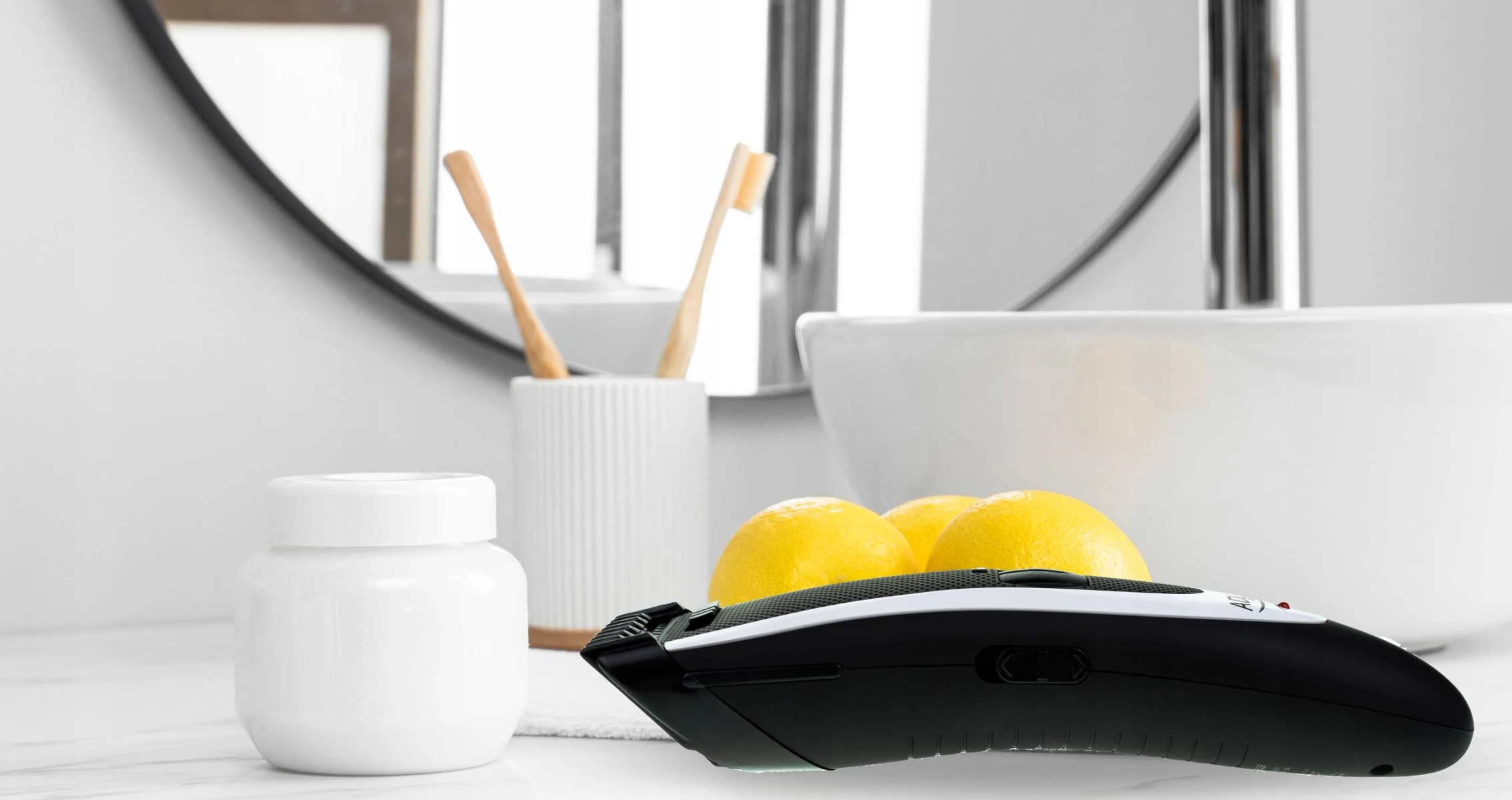 Escena de baño con recortadora, tarro, limones, cepillos de dientes y espejo. La recortadora es blanca y negra.