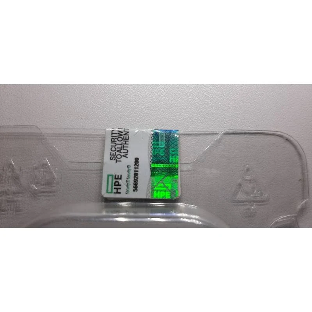 Naklejka zabezpieczająca HPE. Zawiera tekst i wzór holograficzny. Znajduje się na przezroczystym plastiku.