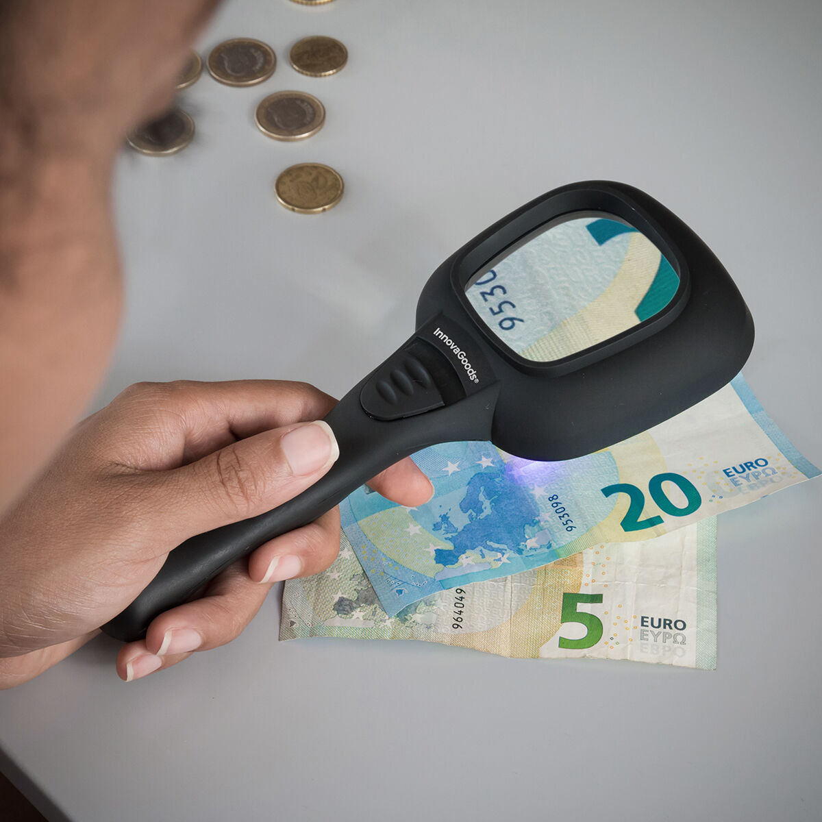 Una persona sostiene una lupa sobre un billete de 20 y uno de 5 euros, con algunas monedas a la vista.