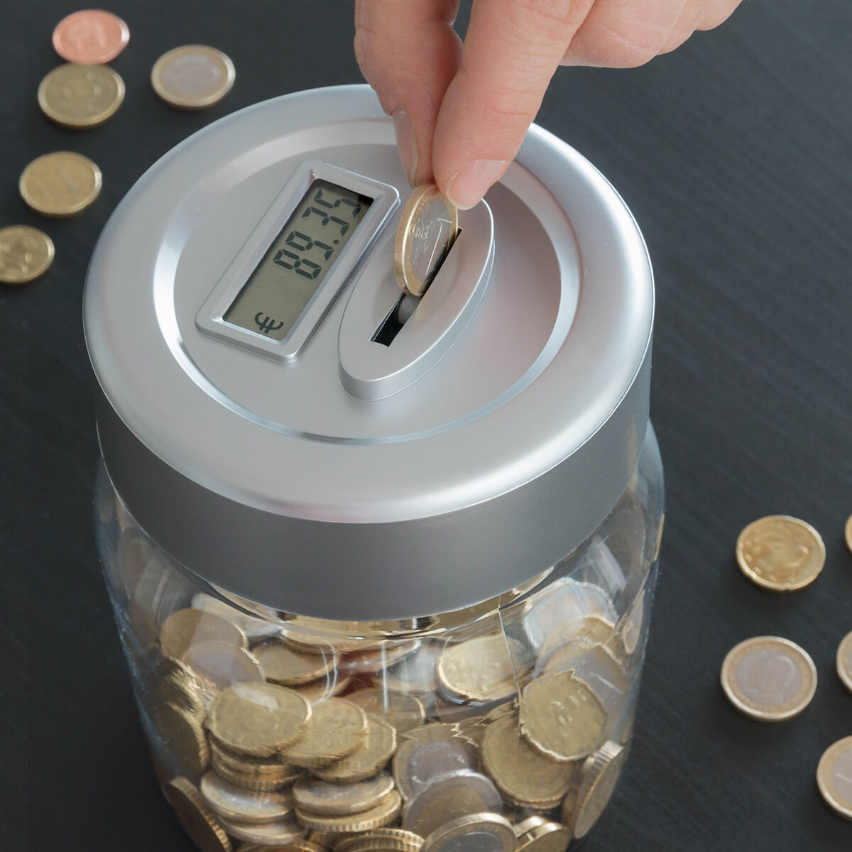 Una persona insertando una moneda de euro en una hucha plateada con forma de tarro. Se ven monedas dentro.