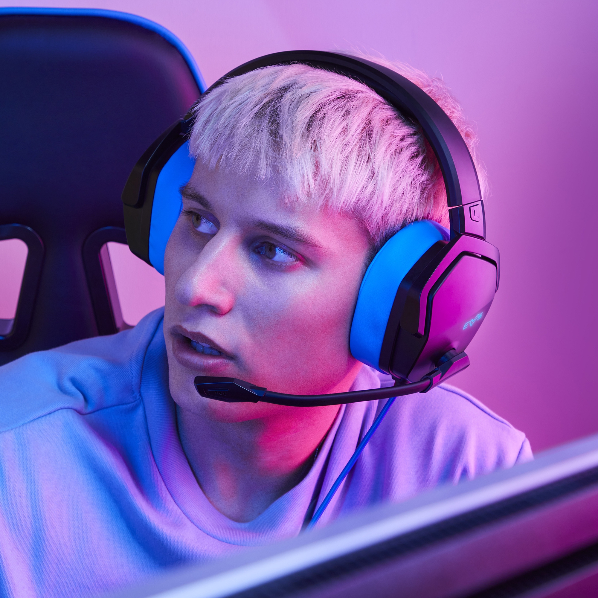 Młody mężczyzna z niebieskimi słuchawkami. Różowo-fioletowe tło. Skupiony wyraz twarzy.