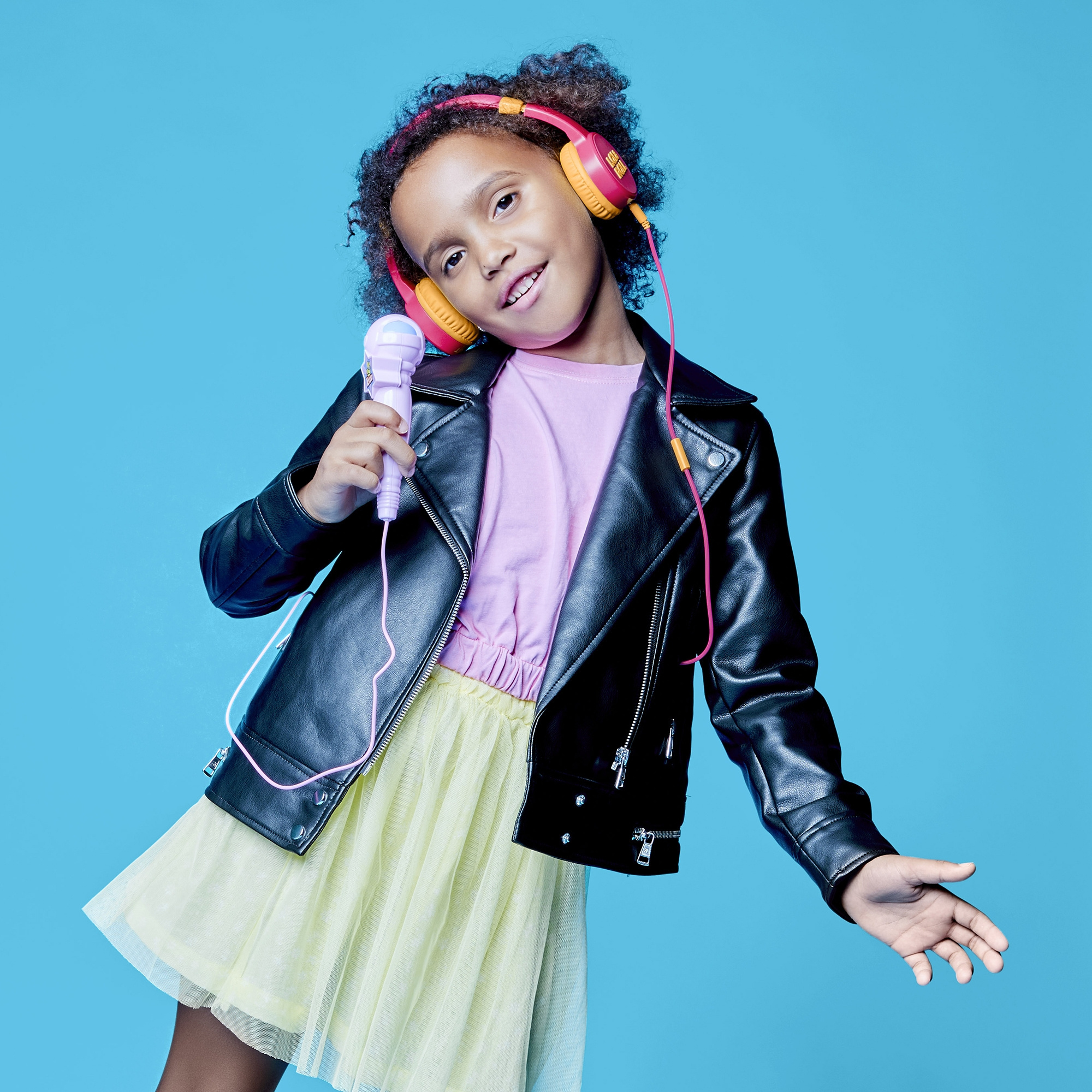 Niña con auriculares y micrófono. Lleva chaqueta negra y falda amarilla, sobre fondo azul.