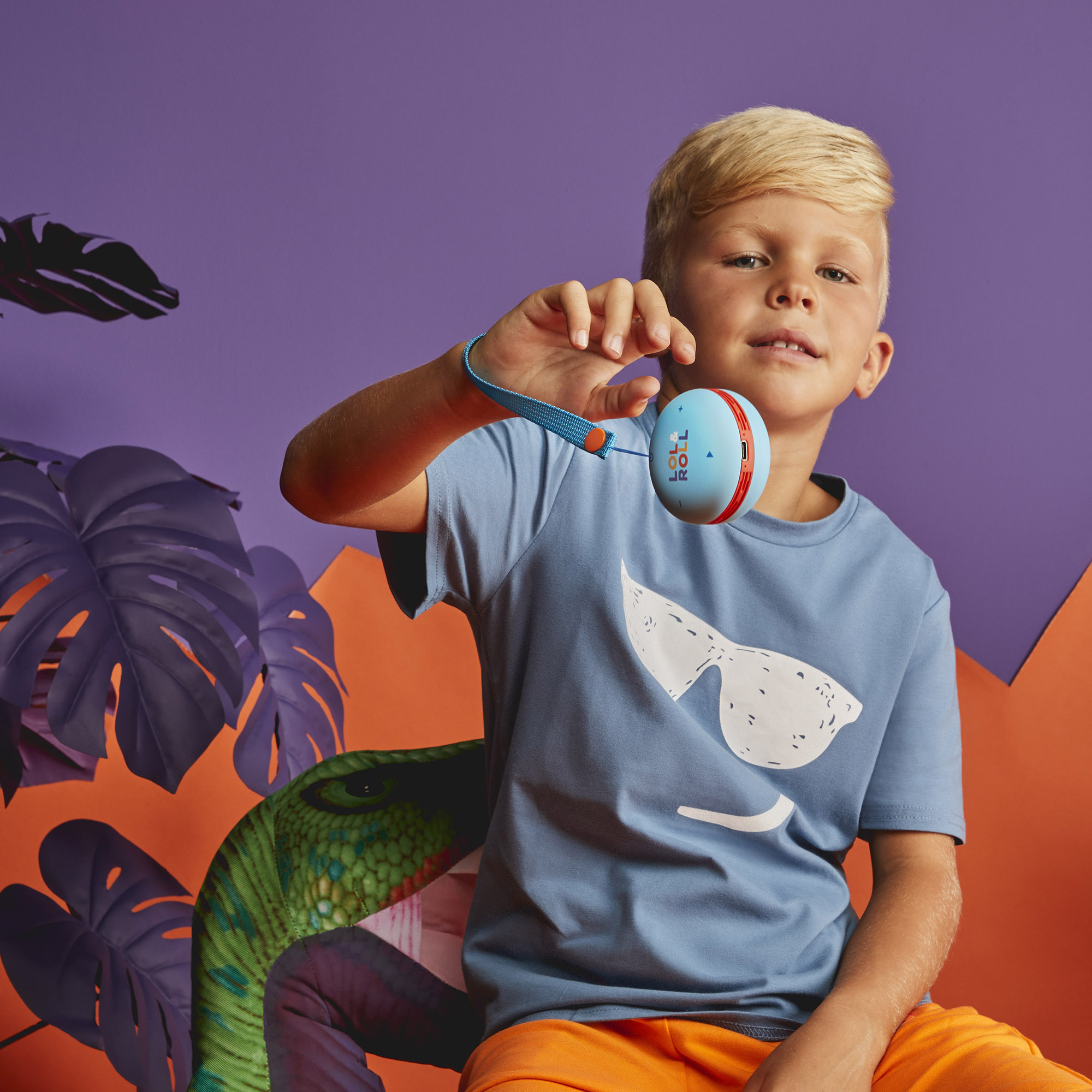 Niño sosteniendo un juguete, camisa azul con diseño de gafas de sol, fondo morado.