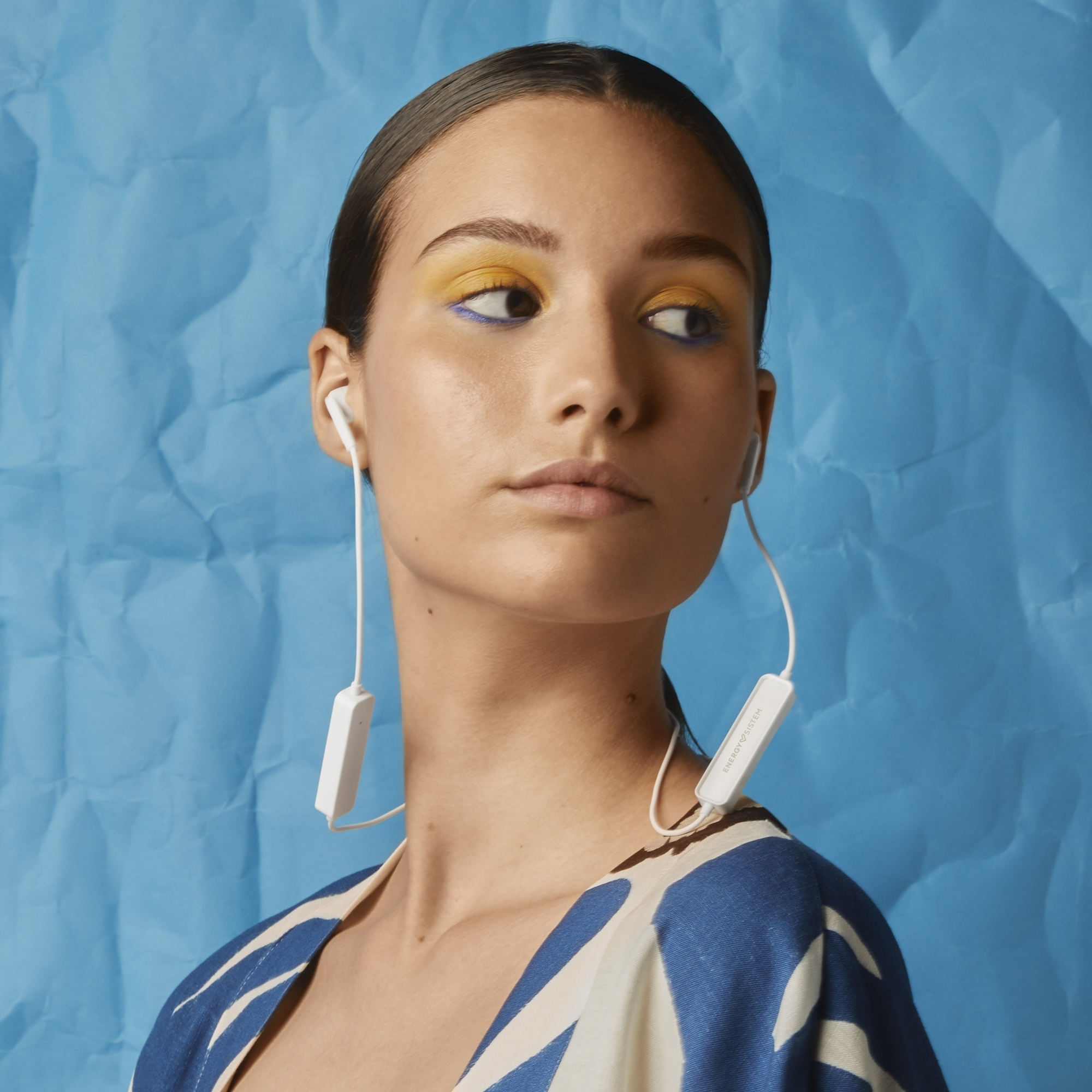 Una donna con ombretto giallo e eyeliner blu indossa auricolari bianchi e un vestito fantasia.