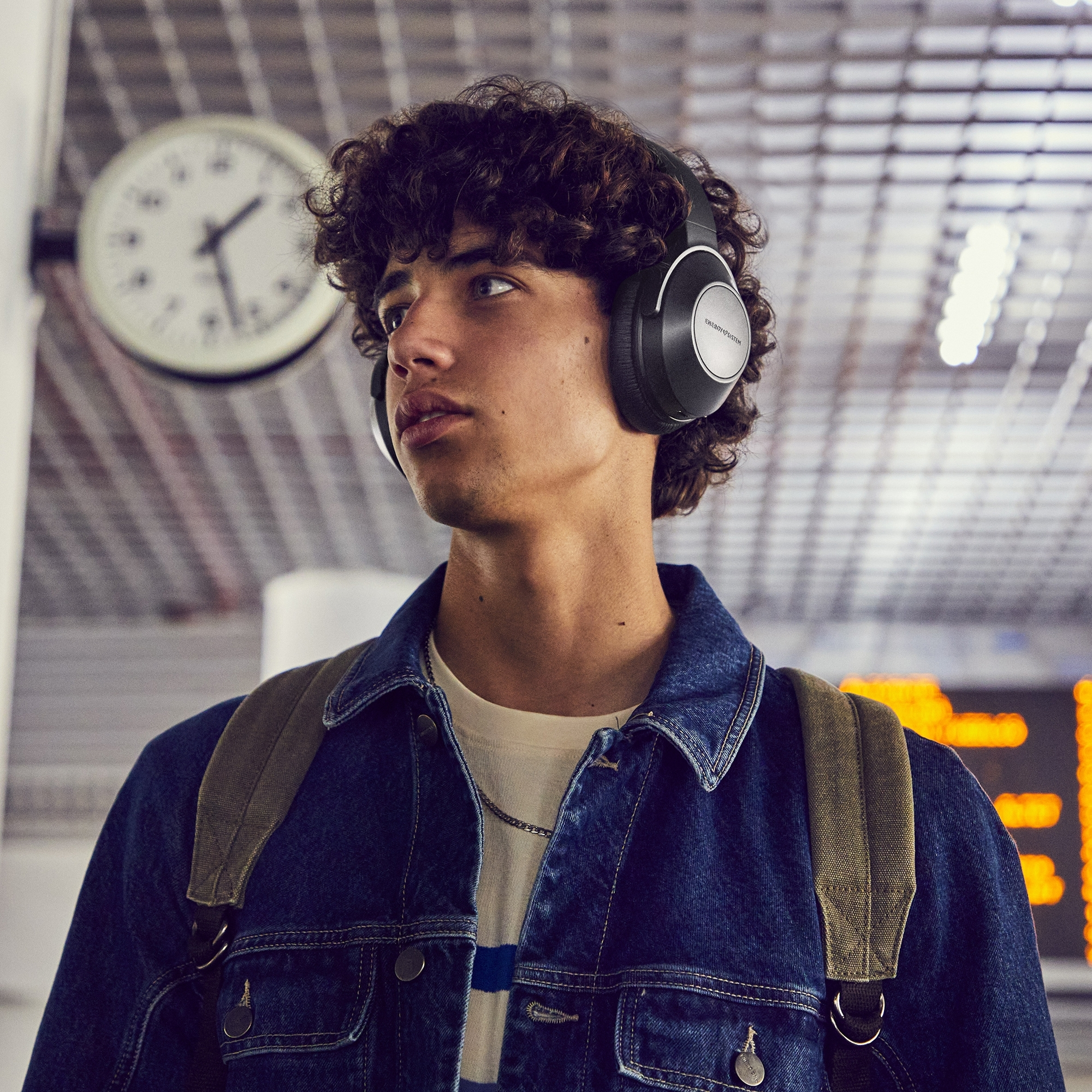 Joven con auriculares negros, mirando hacia otro lado, en una estación de tren.
